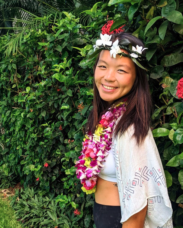 A woman wearing a floral lei and a flower crown, standing in front of lush green foliage with pink flowers, smiling at the camera.