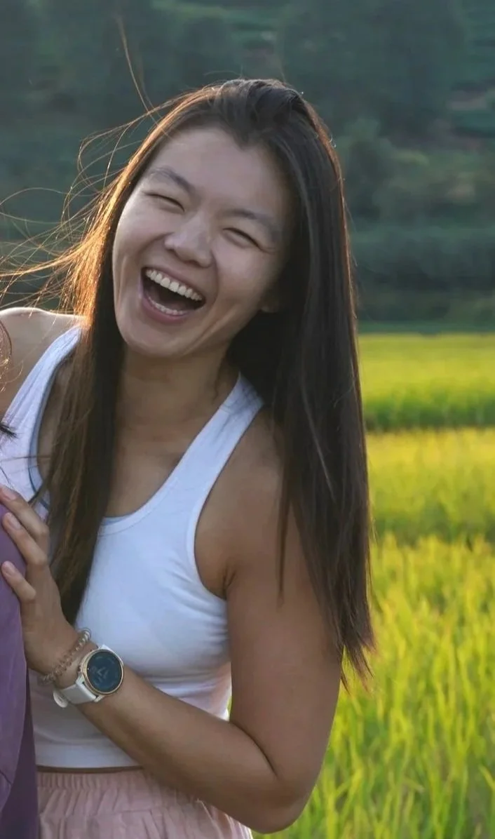 A woman with long dark hair smiling and laughing outdoors in a grassy field during daytime.