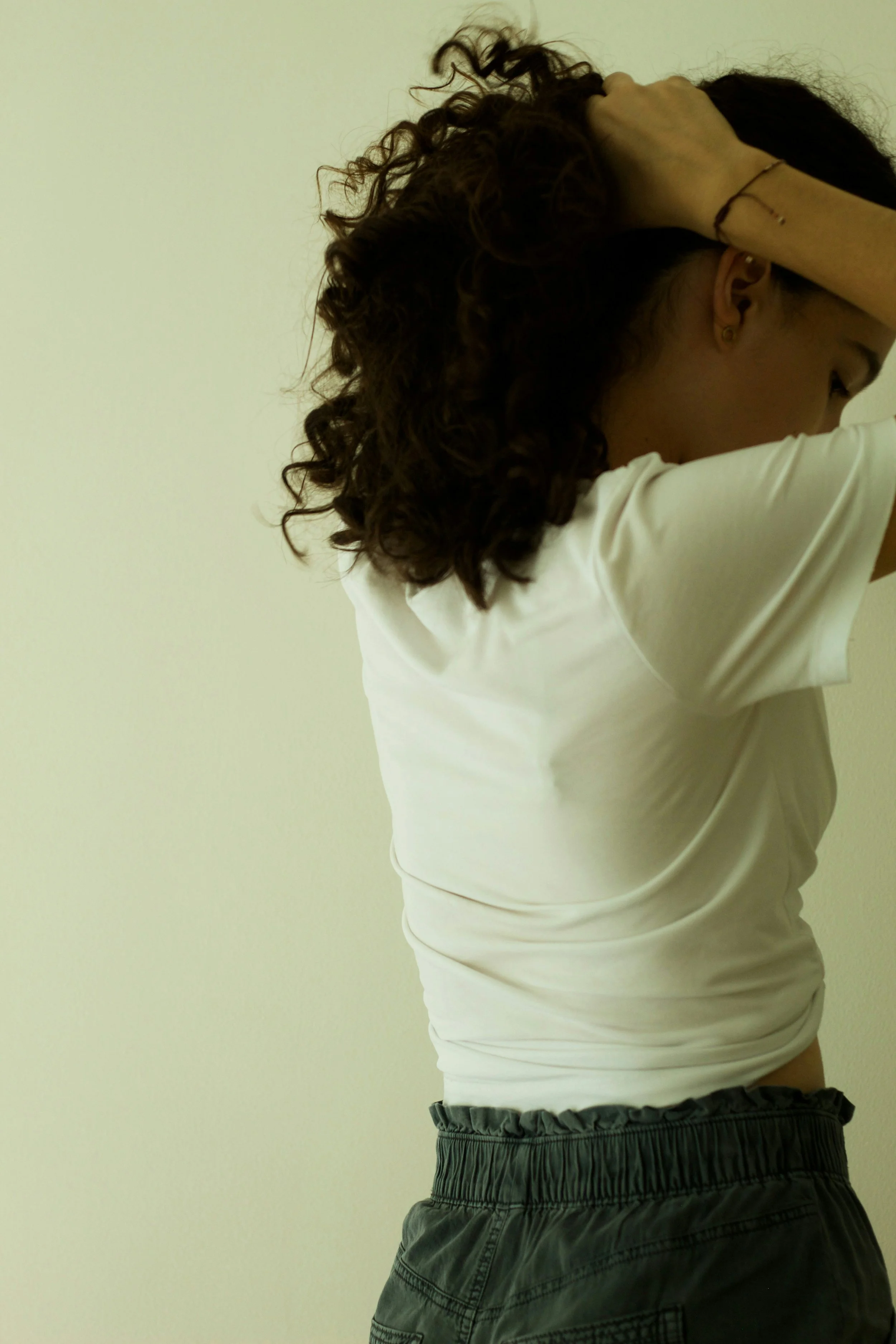A woman with curly dark hair wearing a white t-shirt and dark shorts, holding her head with one hand, against a plain light-colored wall.