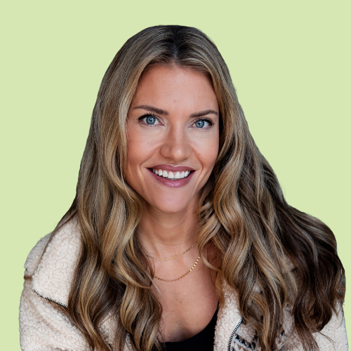 Portrait of a woman with long blonde hair, wearing a beige blazer and black top, smiling with arms crossed against a green background.