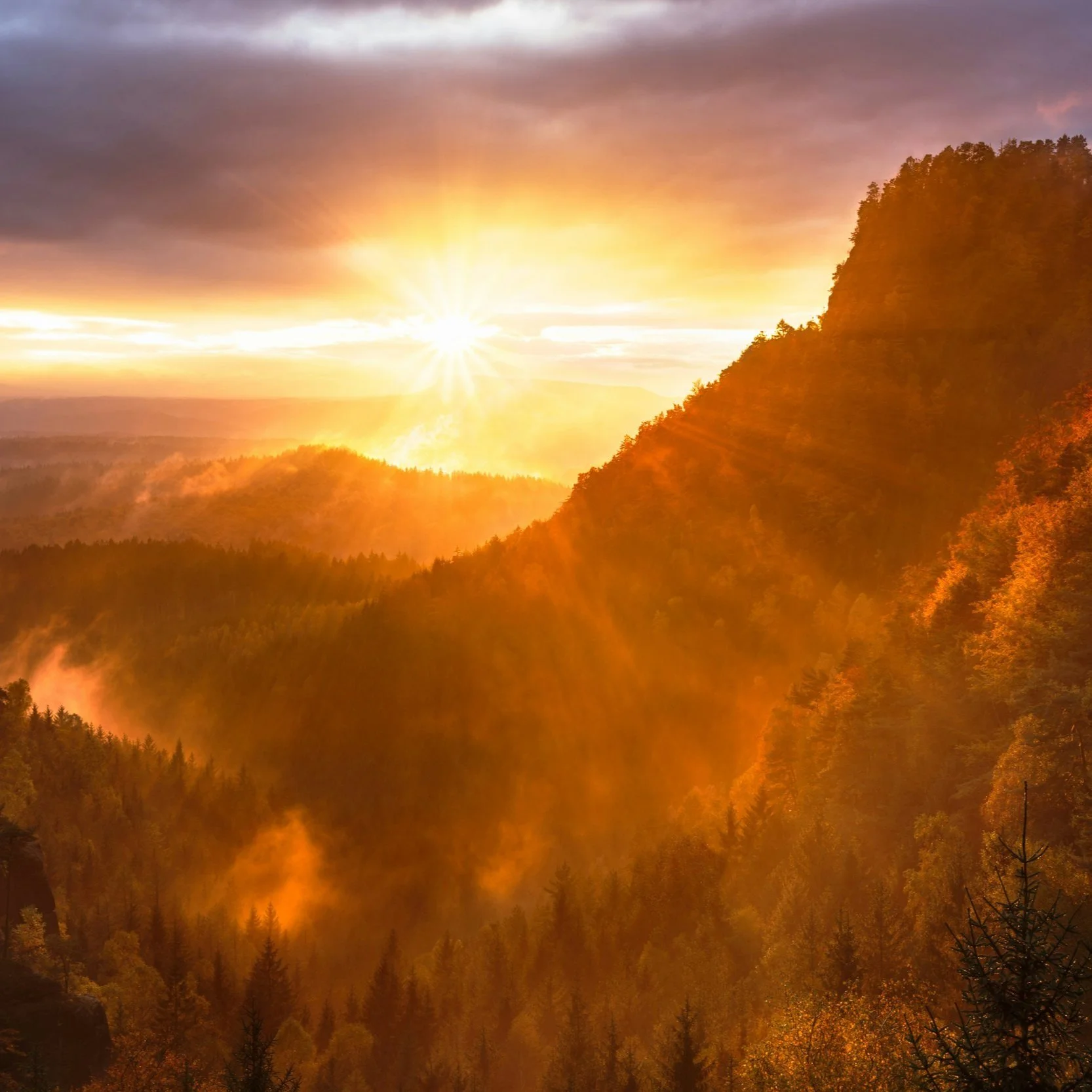 Sunrise over a mountain landscape with a forested valley, rays of sunlight, and orange and purple clouds.