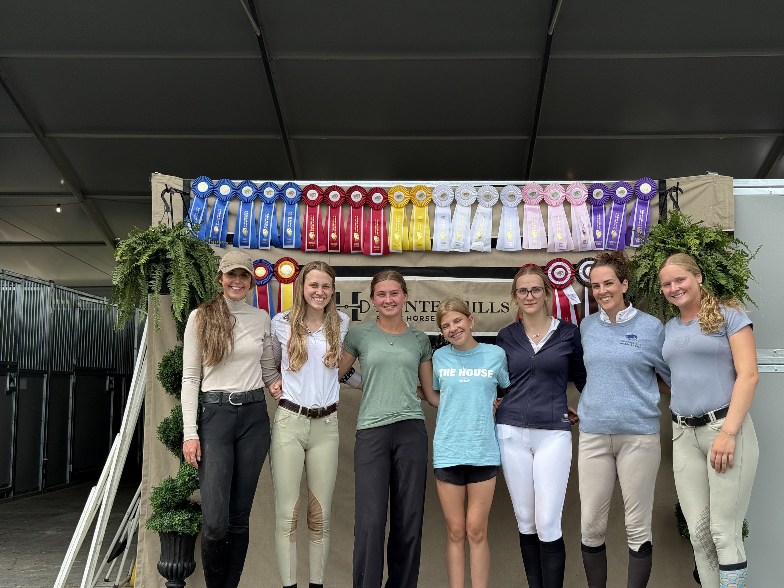Group of young women standing in front of a display of colorful ribbons at a horse competition event, smiling at the camera.