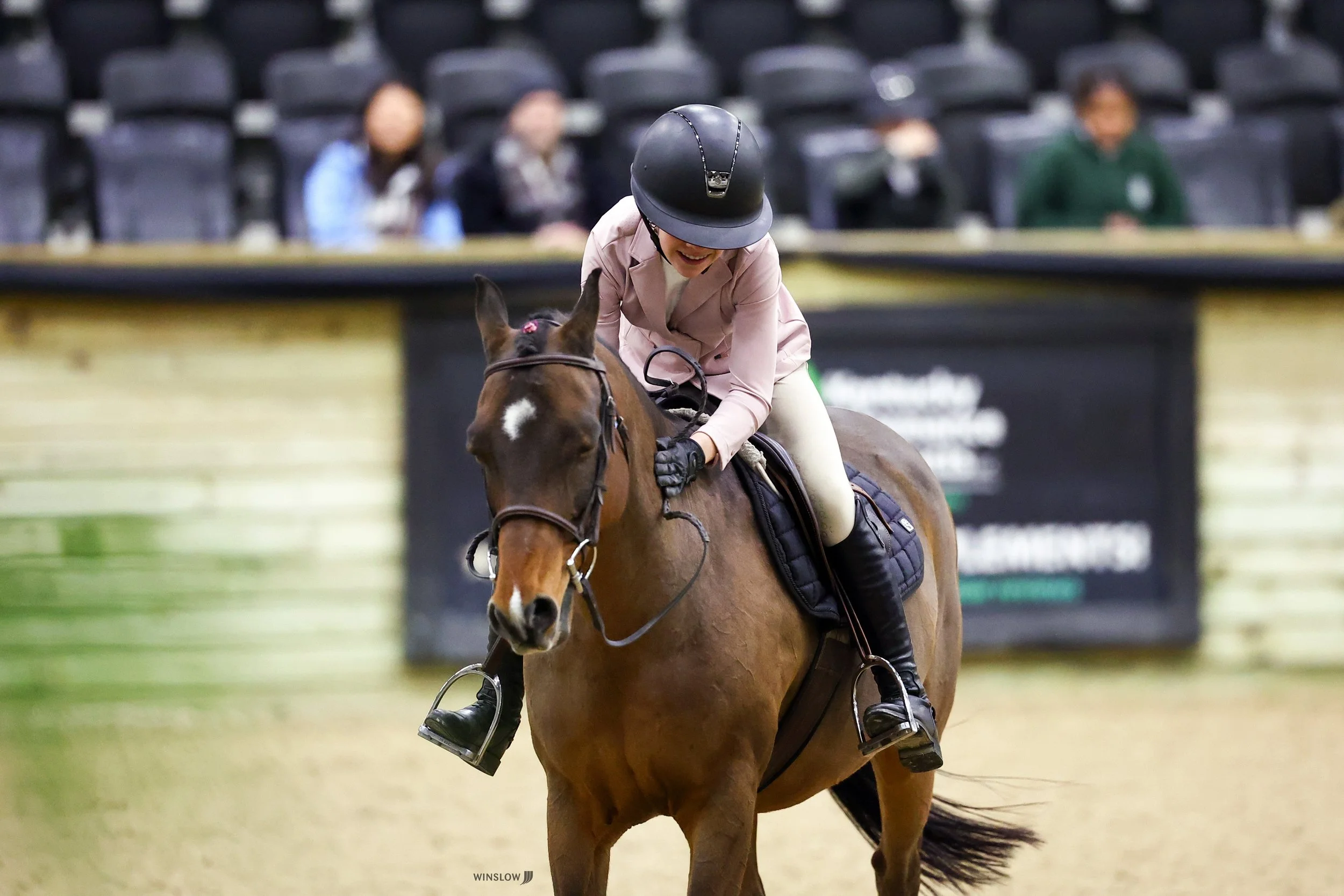 A woman riding a horse in an indoor arena, wearing a helmet and smiling, with a few people sitting in the background.