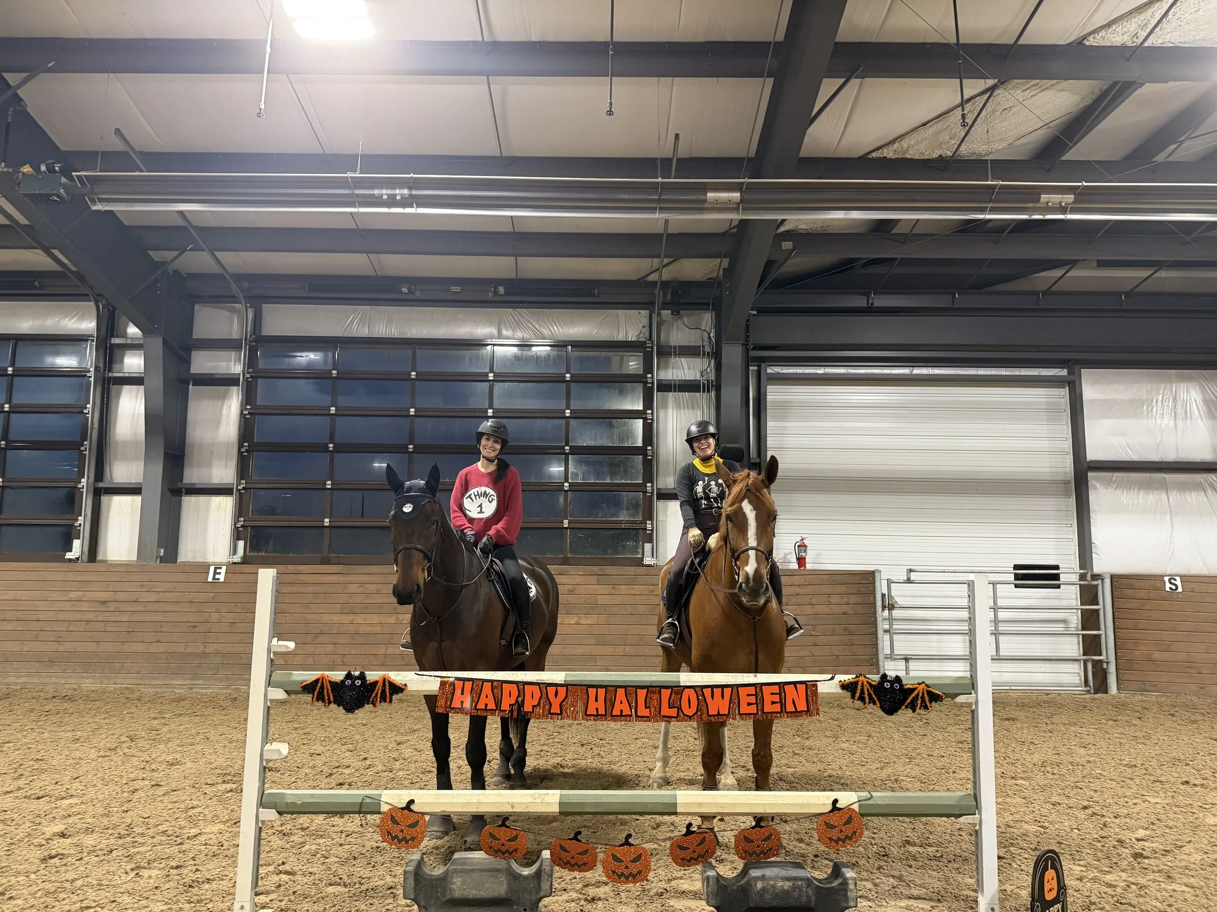 Two women riding horses in an indoor riding arena decorated for Halloween with a banner reading 'Happy Halloween' and black and orange pumpkin decorations.