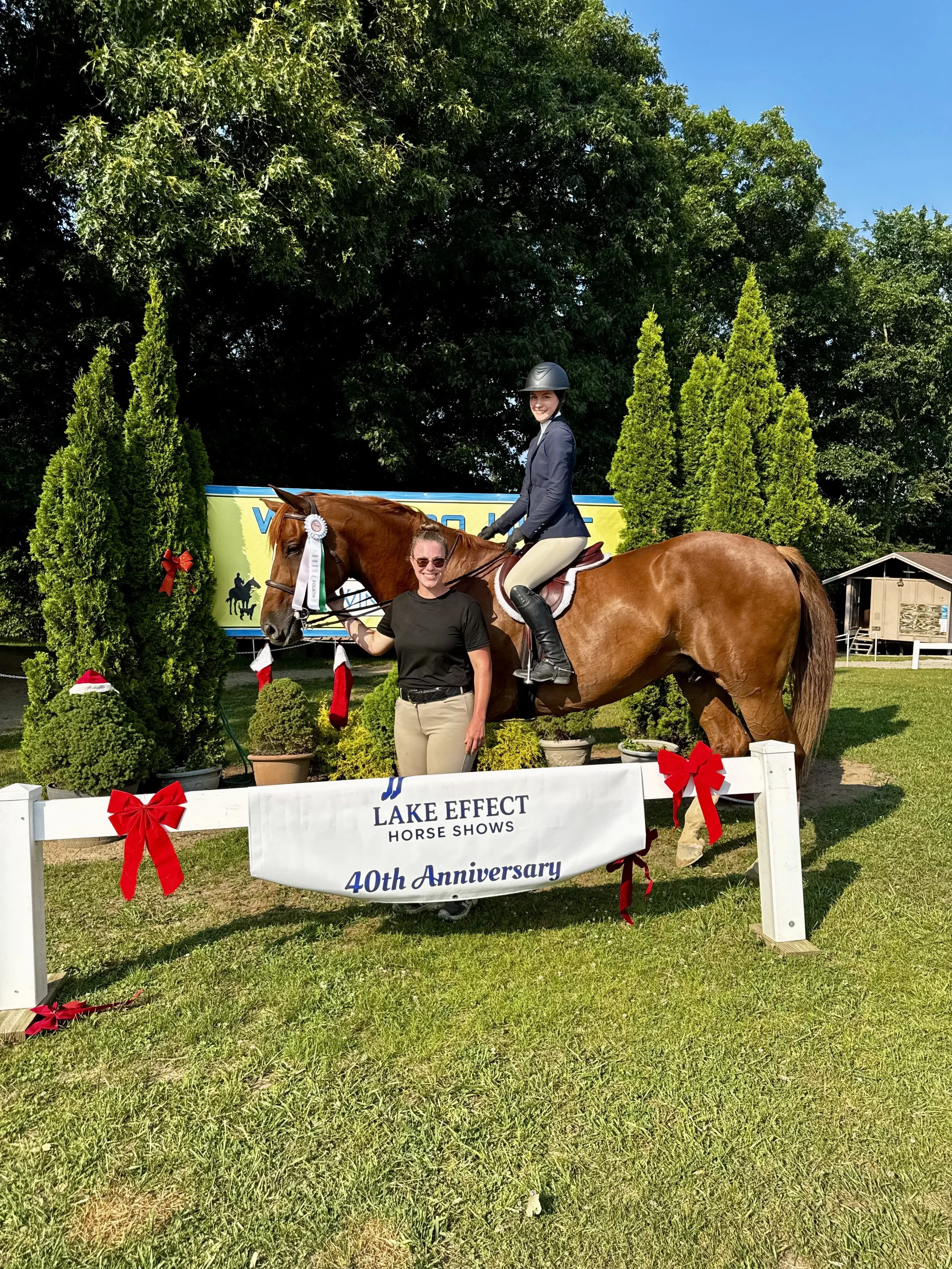 A woman in riding attire riding a chestnut horse at a horse show, with a woman in casual attire standing beside the horse holding the reins. The scene is decorated with small trees, red bows, and a sign that reads "Lake Effect Horse Shows 40th Annive