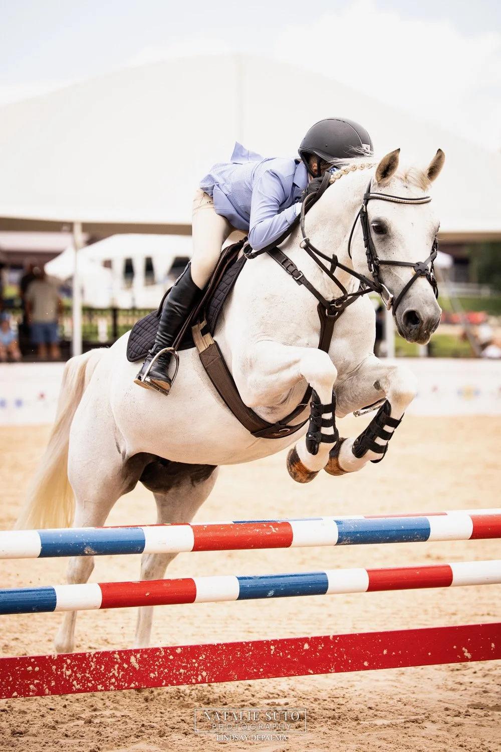 A young female equestrian wearing a helmet and riding jacket jumps over a hurdle on a white horse during a show jumping competition, with spectators and tents in the background.