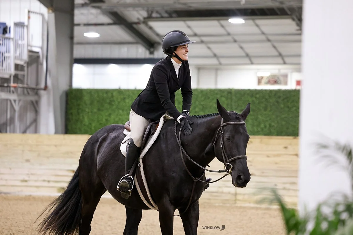 A woman in riding attire, wearing a helmet, riding a black horse in an indoor riding arena with a wooden fence and green wall in the background.