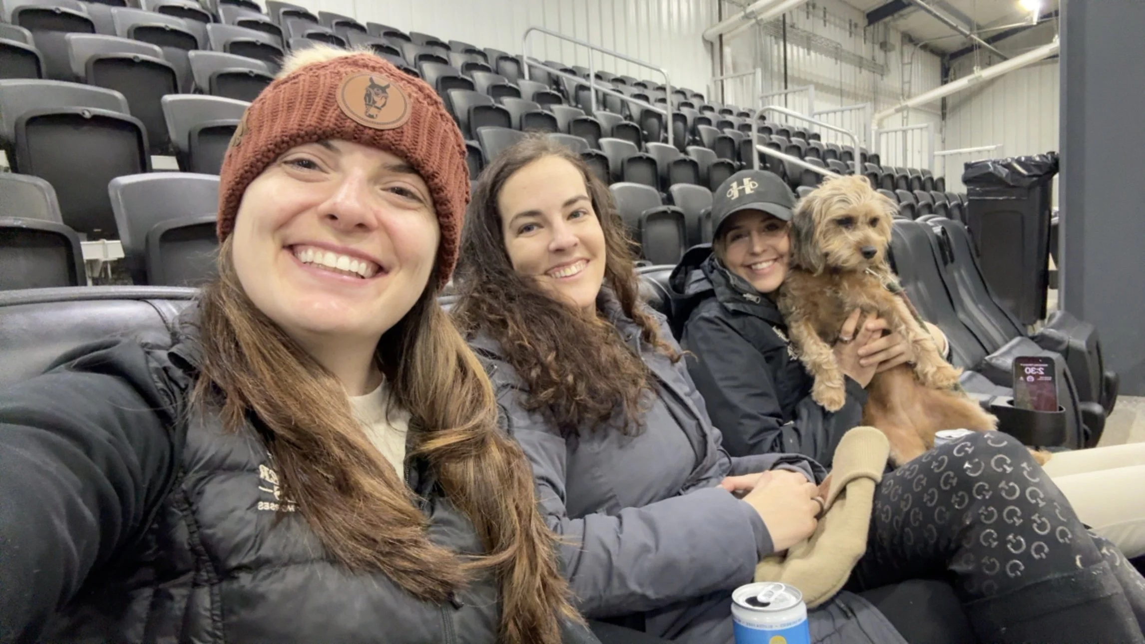 Four women and a small dog sitting in stadium seats, smiling at the camera, with black empty seats behind them.