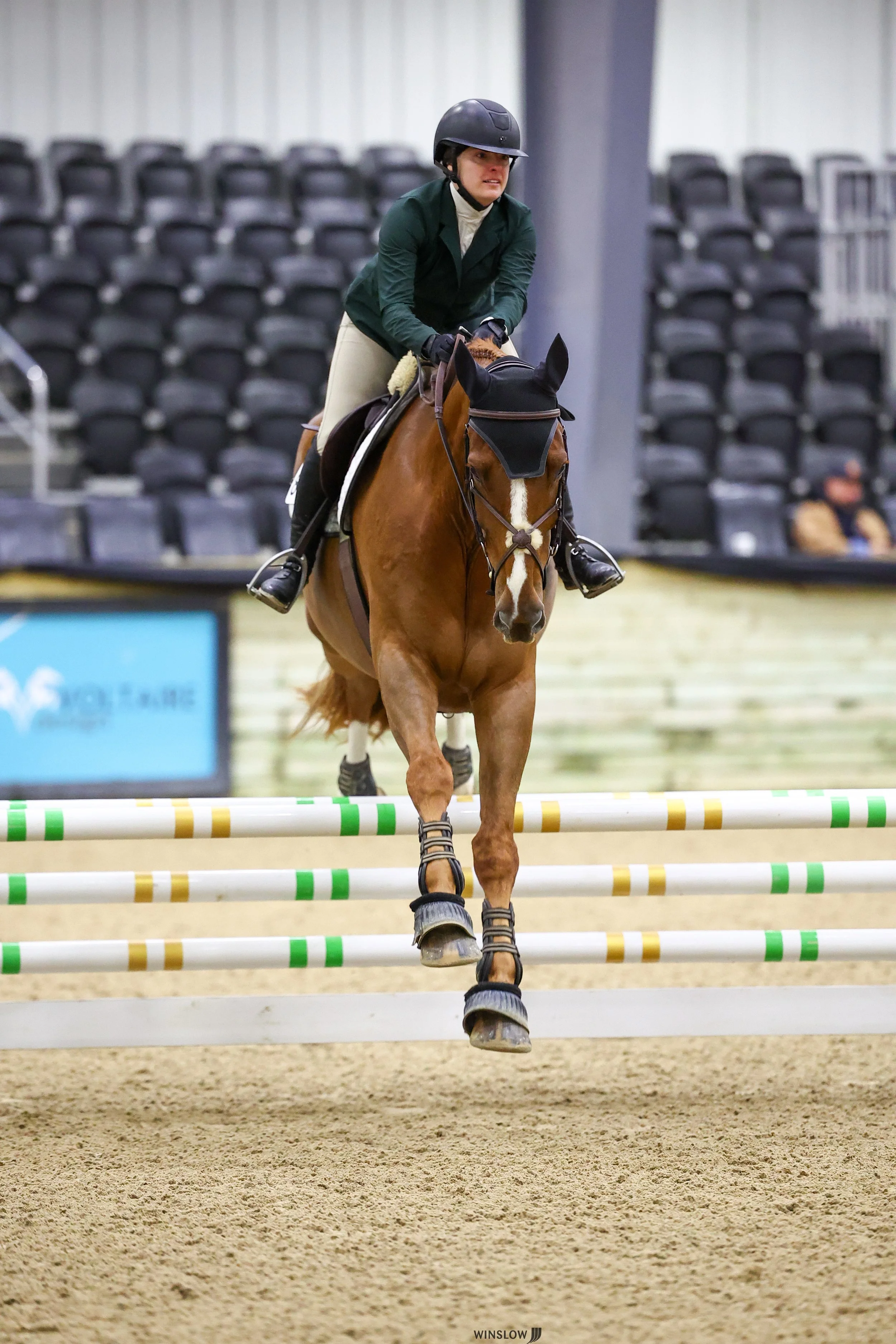 A female equestrian athlete riding a brown horse over a show jumping obstacle with colorful poles in an indoor arena.