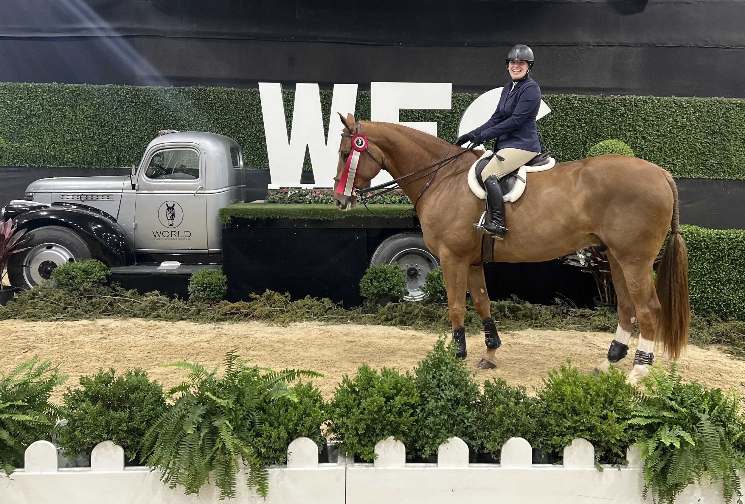A person riding a horse at an equestrian event, smiling, wearing a helmet and jacket, with a blue and white rosette ribbon on the horse's bridle. They are on a riding arena with green bushes and ground decorations, behind a truck with the logo 'World
