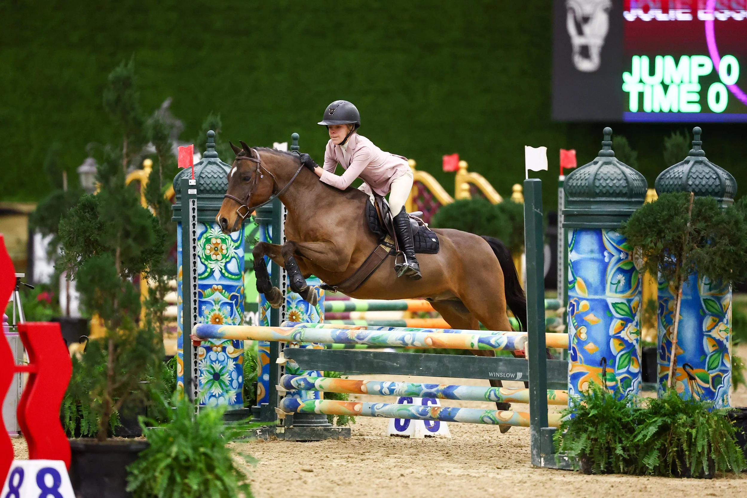 A rider and horse jumping over an obstacle during an equestrian event, with colorful decorative poles and a digital scoreboard in the background.