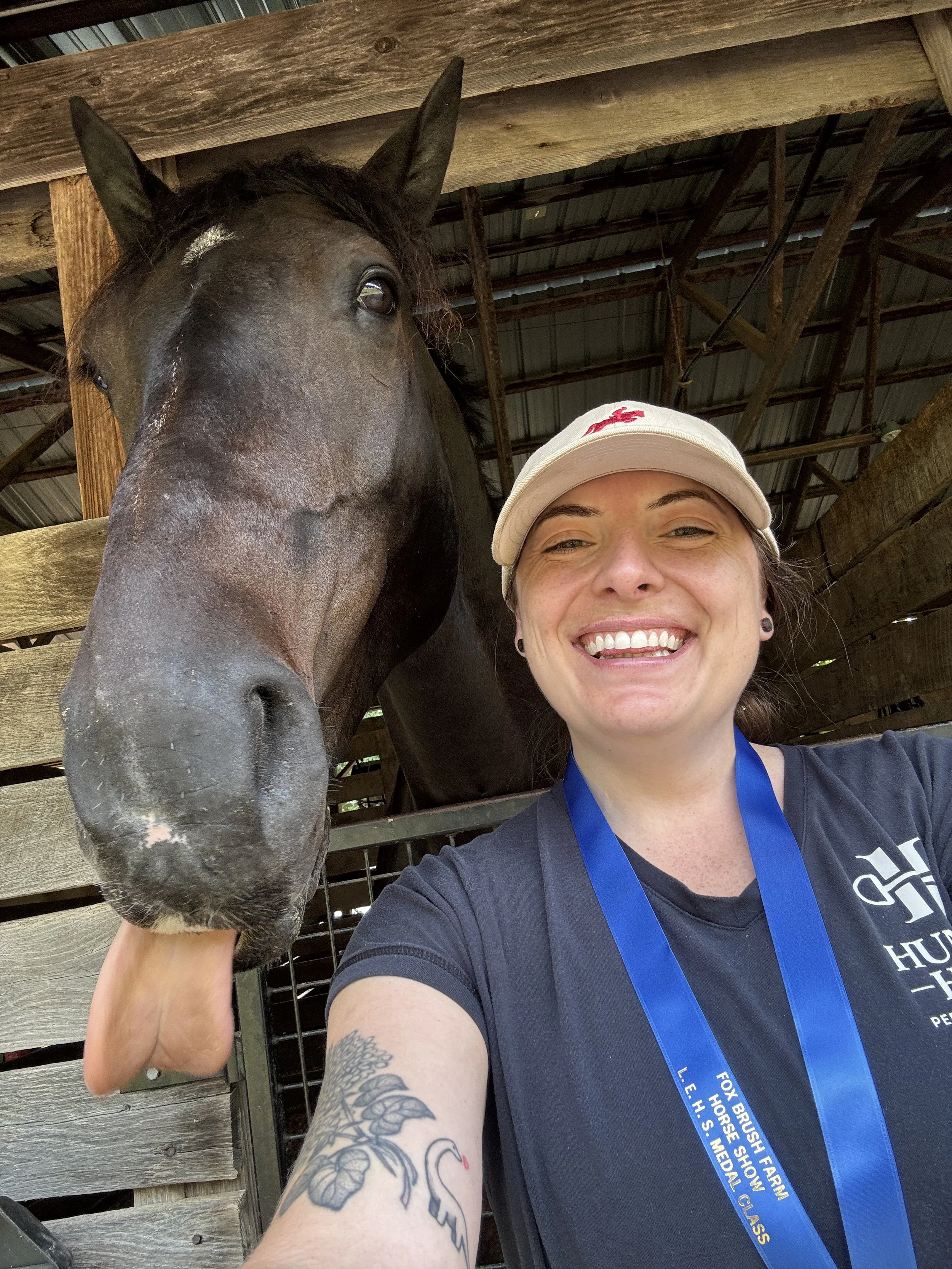 A smiling woman wearing a white baseball cap and a black shirt, taking a selfie with a horse in a wooden stable.