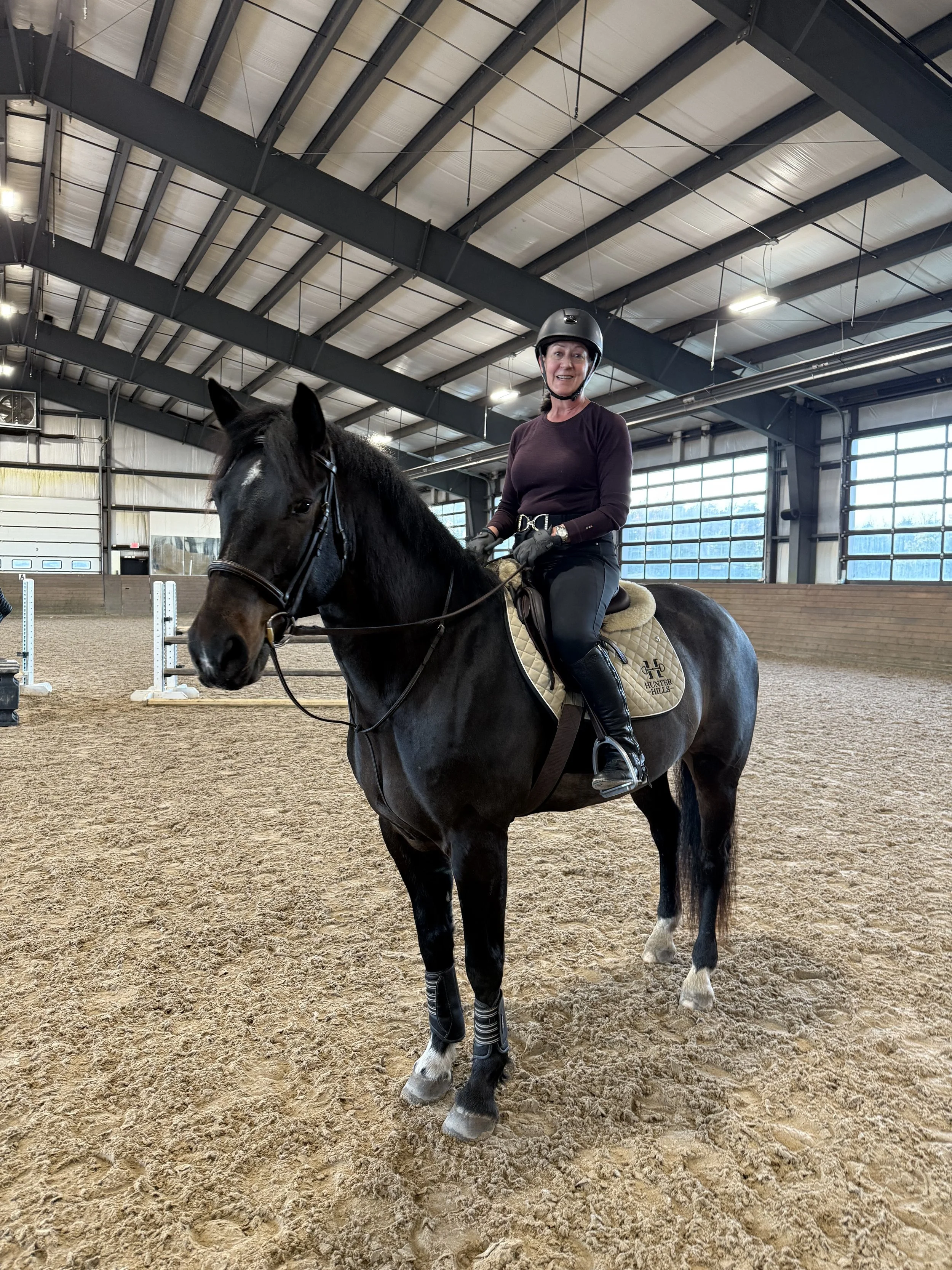 A woman riding a dark brown horse with white markings on its face and socks inside an indoor riding arena. She is wearing a black helmet, gloves, black boots, and a dark long-sleeve shirt. The horse has a beige saddle with a logo, and the arena has a