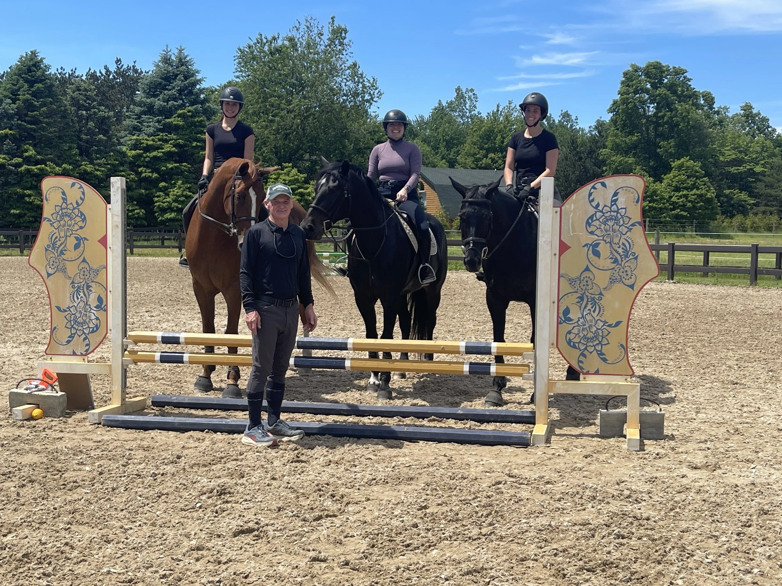 Three women on horseback with helmets, one man standing beside them, in an outdoor riding arena with decorative panels, trees, and a building in the background.