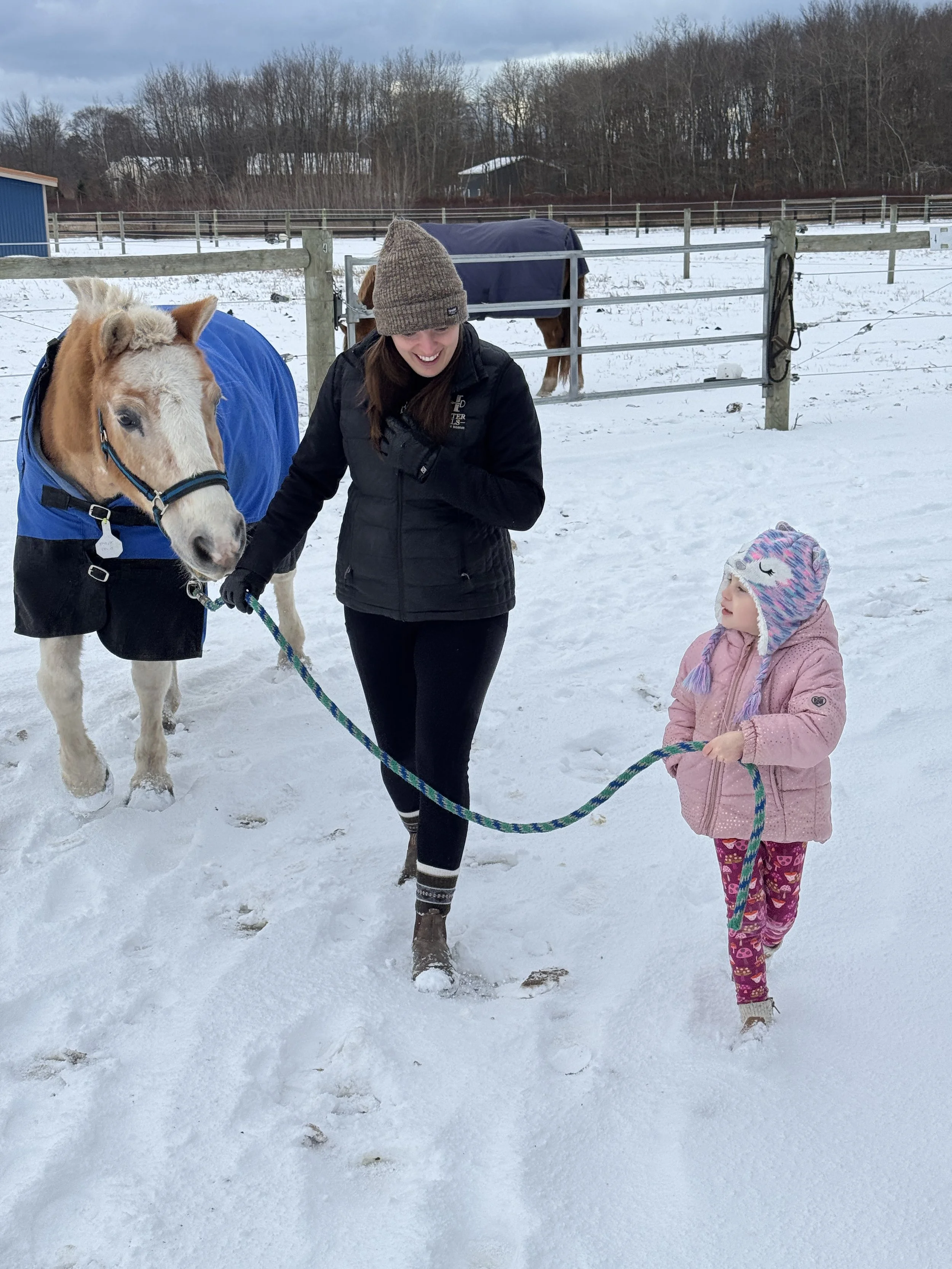 A woman with long brown hair wearing a brown knit hat and black jacket, and a little girl wearing a pink coat and colorful hat, with a horse in a blue blanket and a black blanket behind them in a snow-covered outdoor setting.