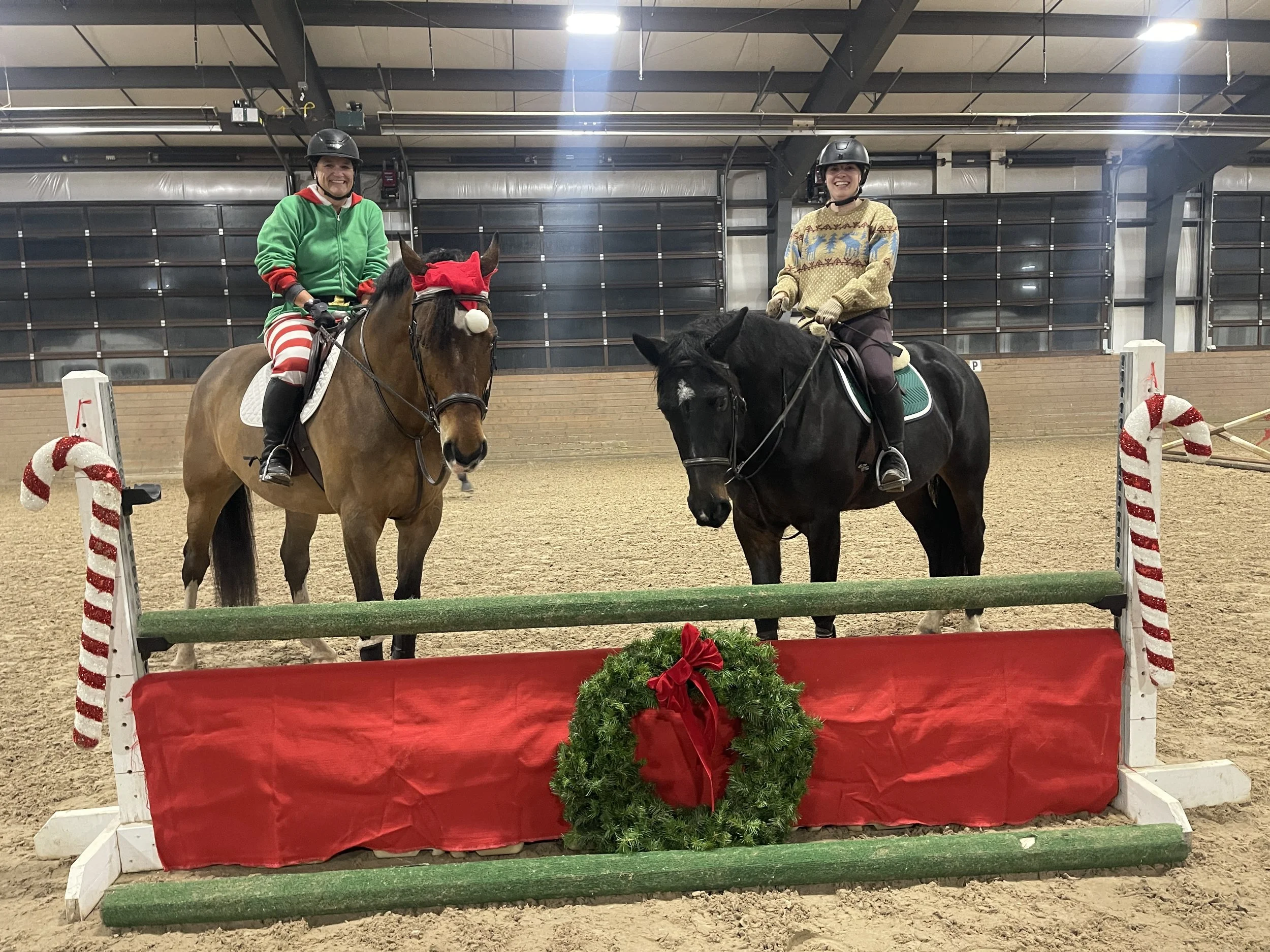 Two women wearing helmets and festive sweaters riding horses inside an indoor equestrian arena decorated for Christmas with a jump obstacle wrapped in red and white candy cane stripes and a holiday wreath. One woman is dressed in a green sweater and 
