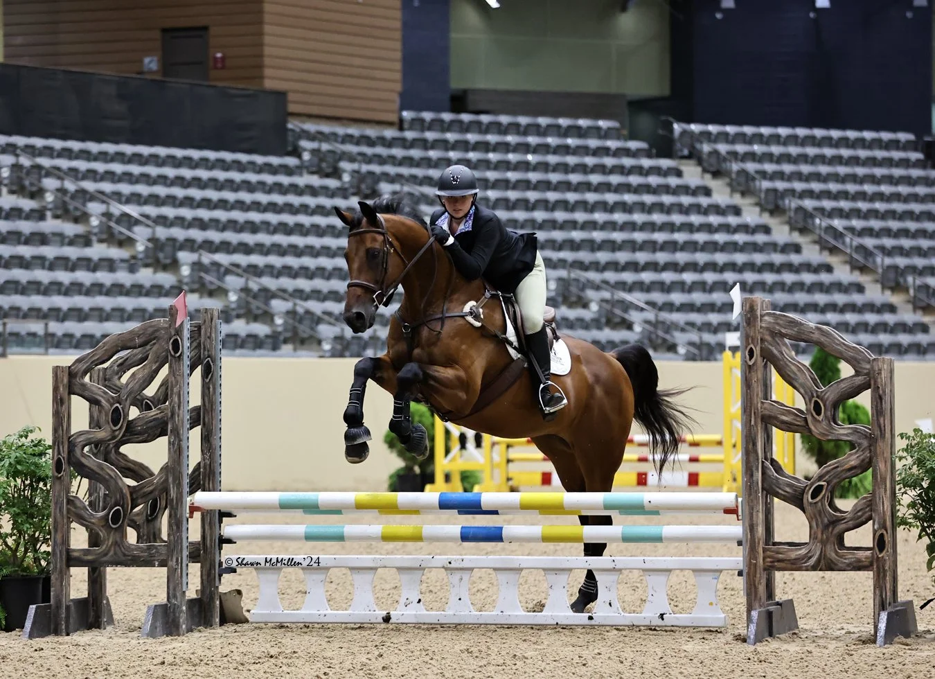 A female equestrian rider in professional riding attire, including a helmet and jacket, guides a brown horse over a colorful show jumping obstacle in an indoor arena with empty stadium seating in the background.
