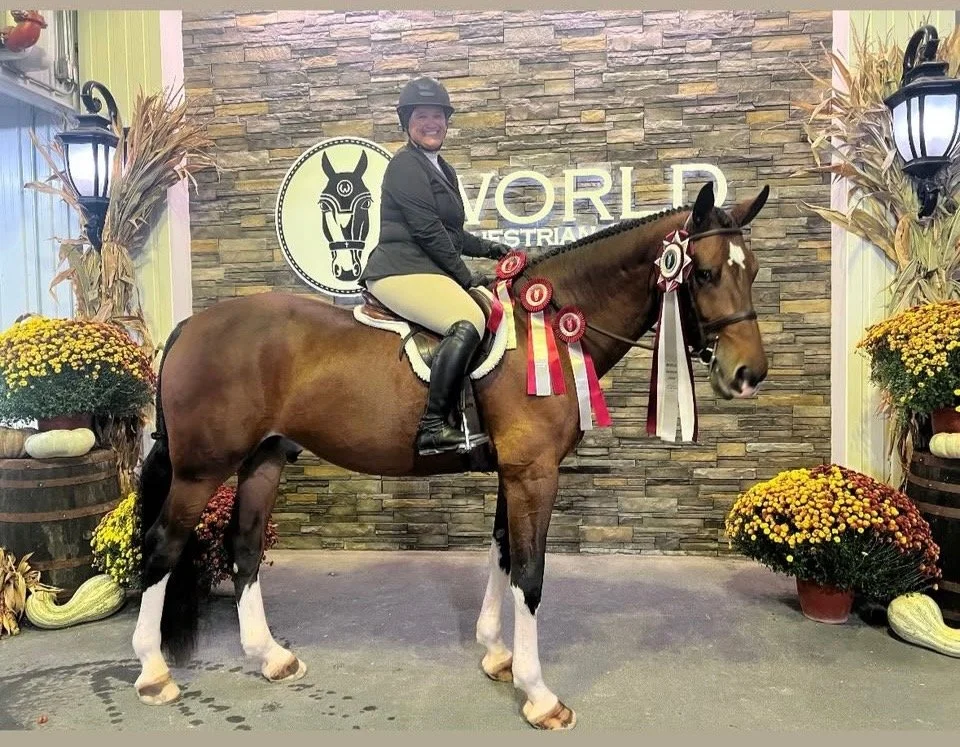 A woman in riding attire sitting on a brown horse decorated with ribbons at an indoor equestrian event. The background has a stone wall with a logo and the word "WORLD" visible. There are potted flowers and pumpkins around the horse.