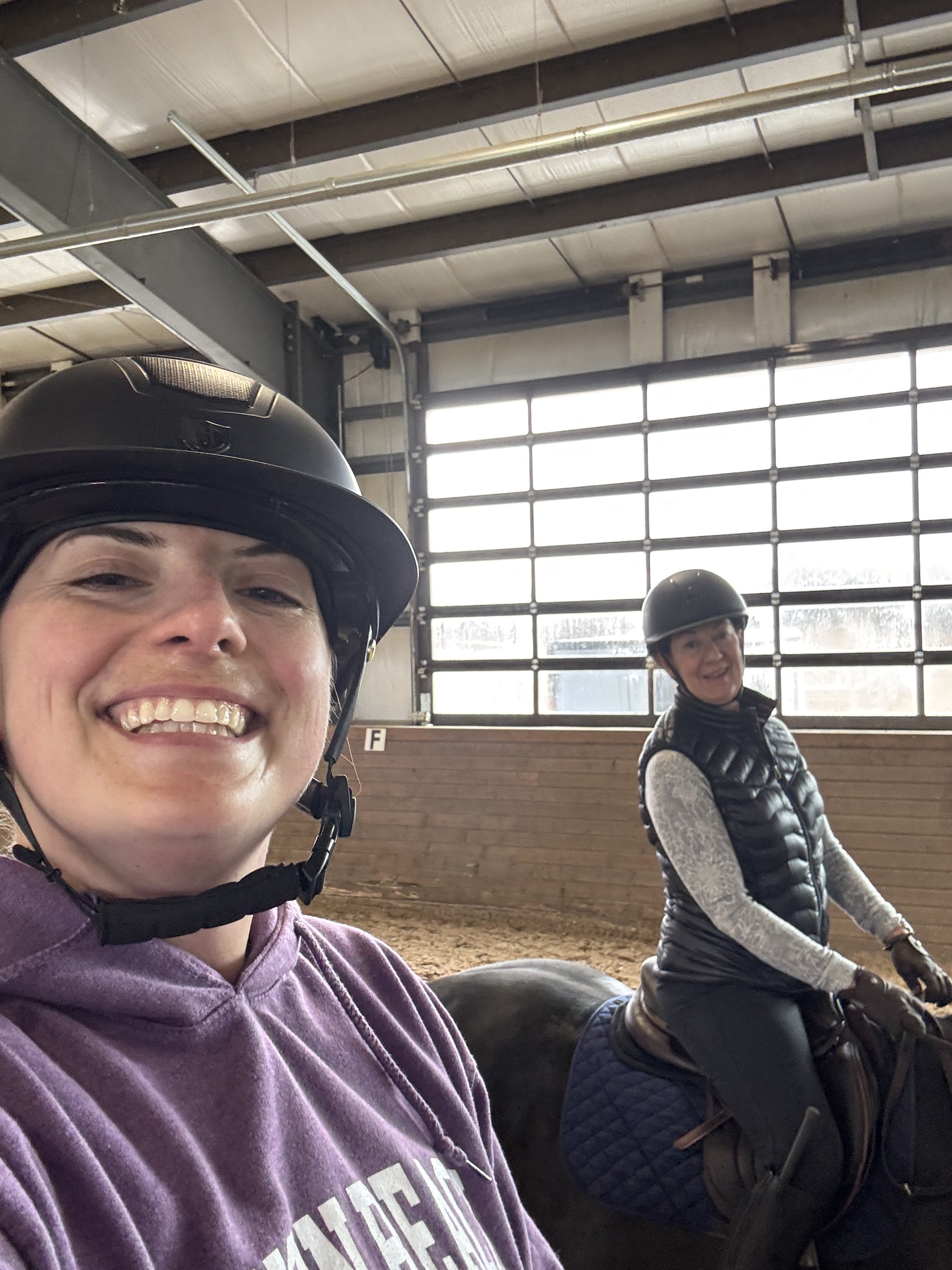Two women wearing helmets and riding gear, sitting on horses inside an indoor riding arena with large windows and metal beams.