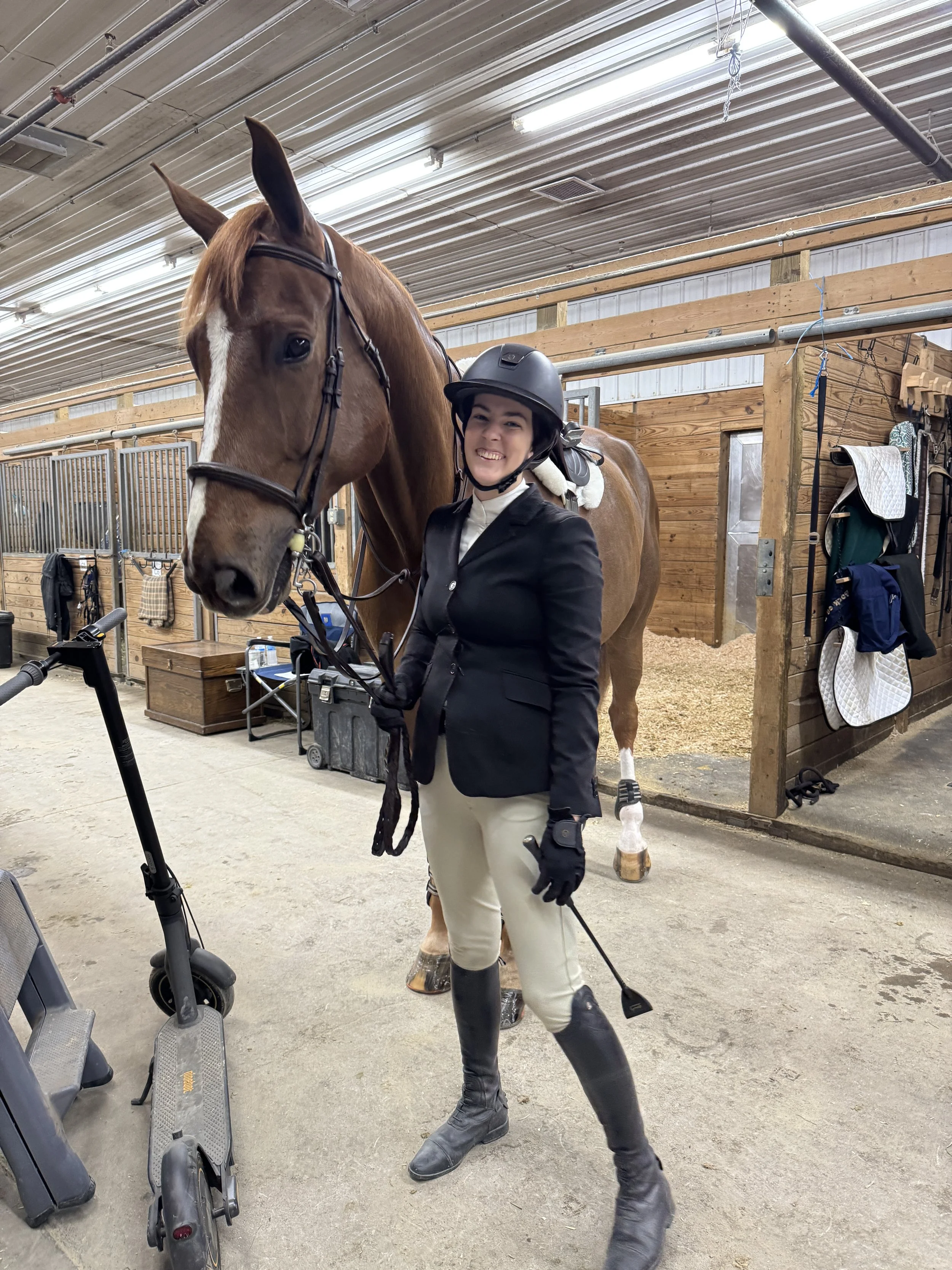 A young woman in riding attire, including helmet and riding boots, stands inside a stable with a brown horse. She is smiling and holding the horse's reins.