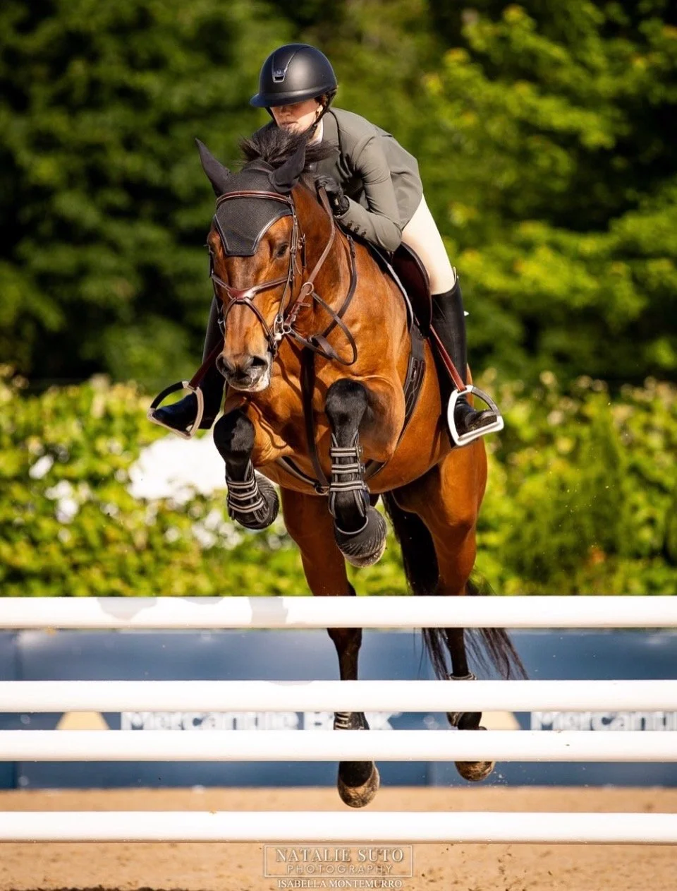 A female equestrian rider in a black helmet and beige riding outfit jumping over a hurdle on a brown horse during a show jumping competition.