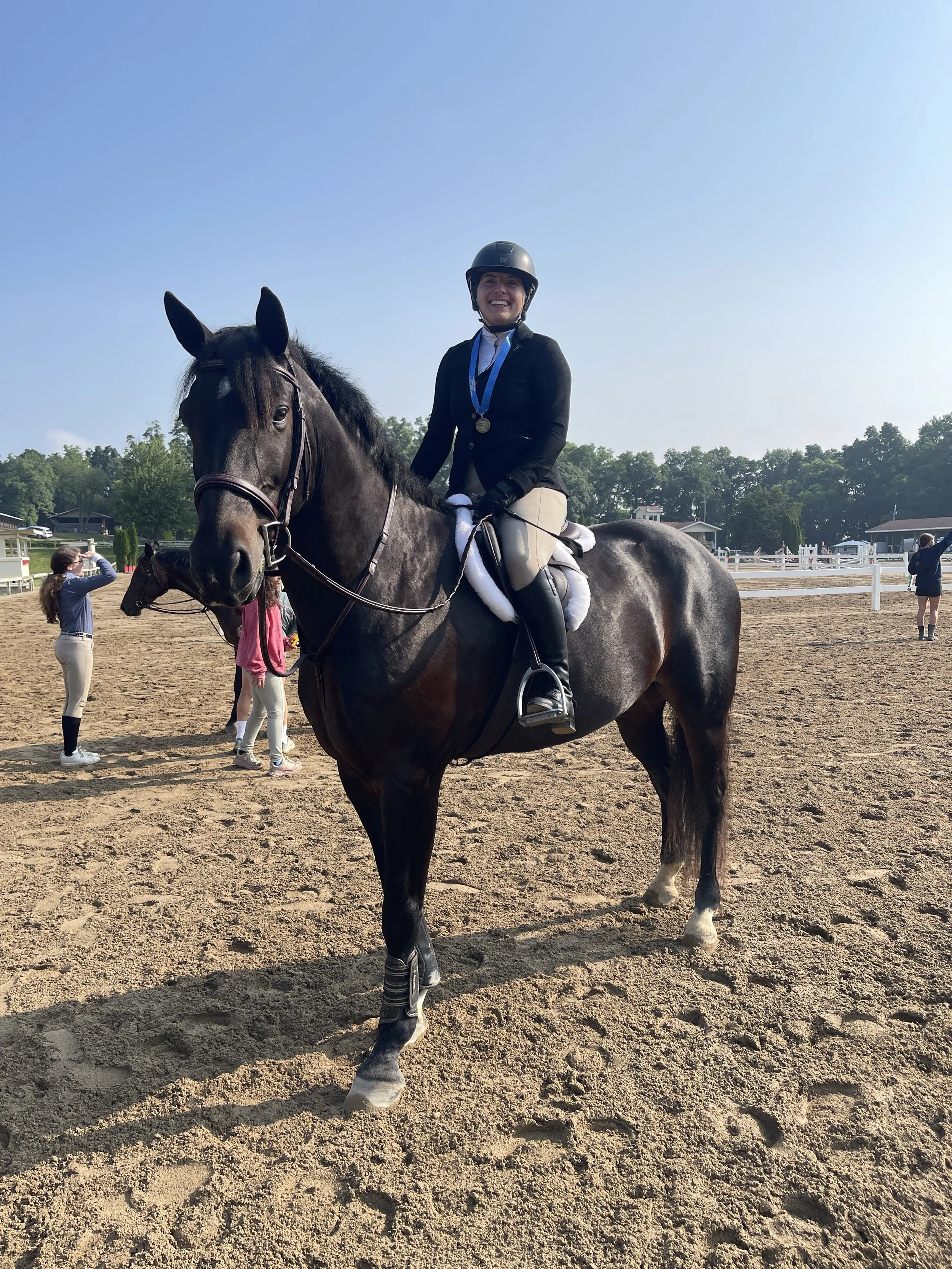A woman in riding gear and a helmet riding a dark horse at an outdoor equestrian arena. She is smiling and has a medal around her neck. There are people and buildings in the background.