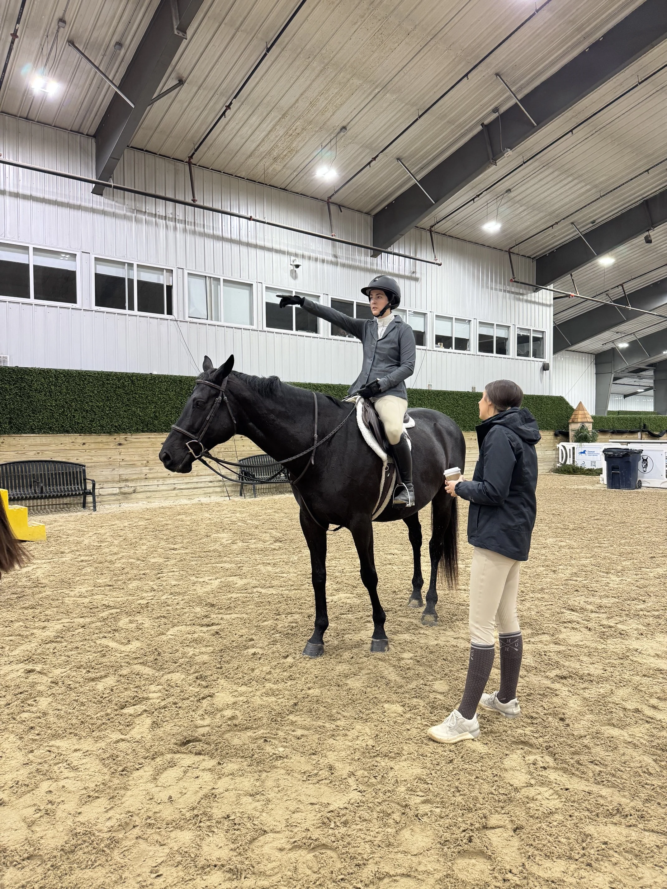 A woman in equestrian attire sitting on a black horse inside an indoor riding arena. A trainer is standing next to the horse, and a person in riding gear is gesturing with their right arm, possibly giving instructions or signaling.