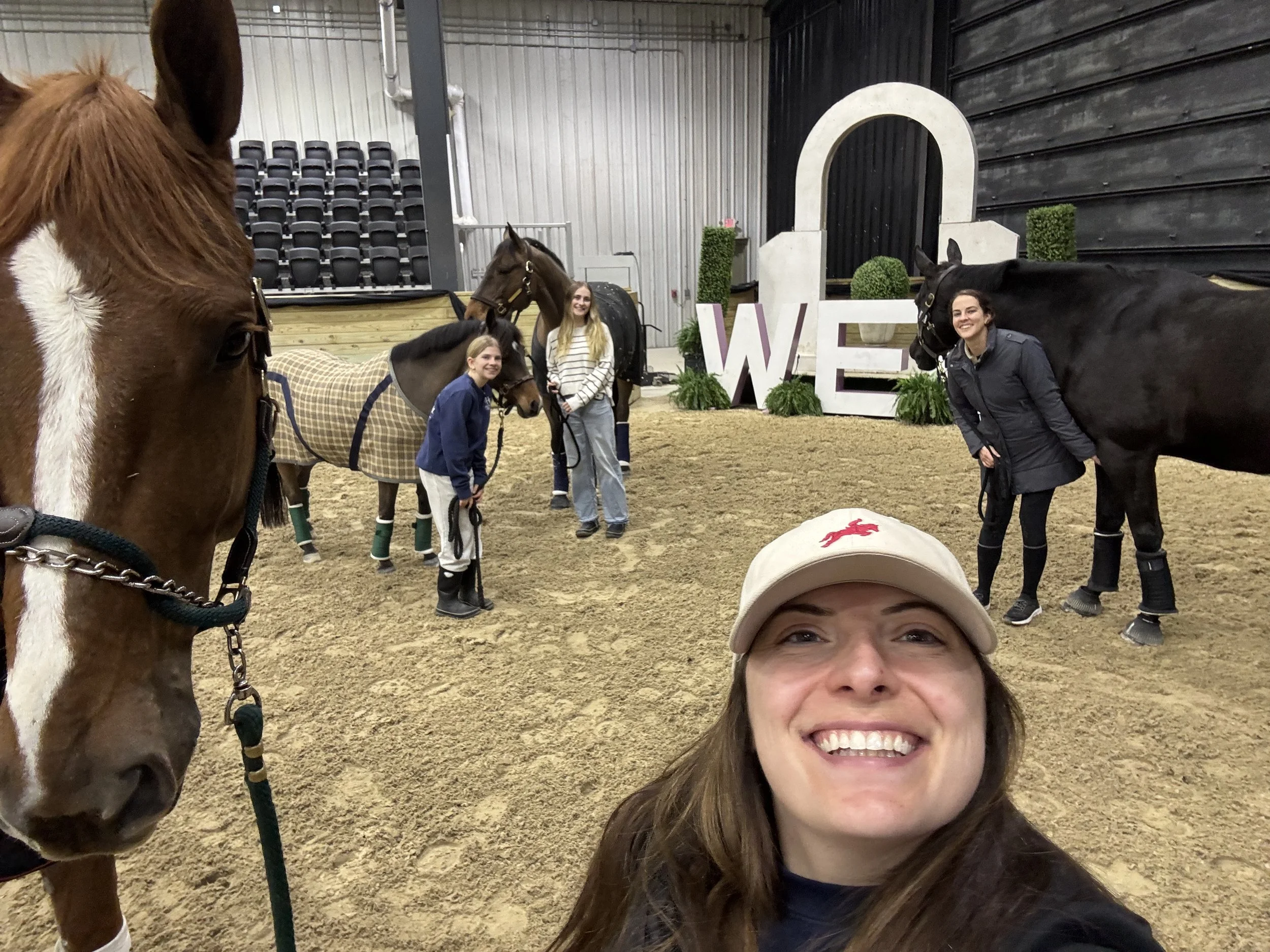 Group of women and horses inside an indoor riding arena with a large white decorative letter 'W' and topiary plants.