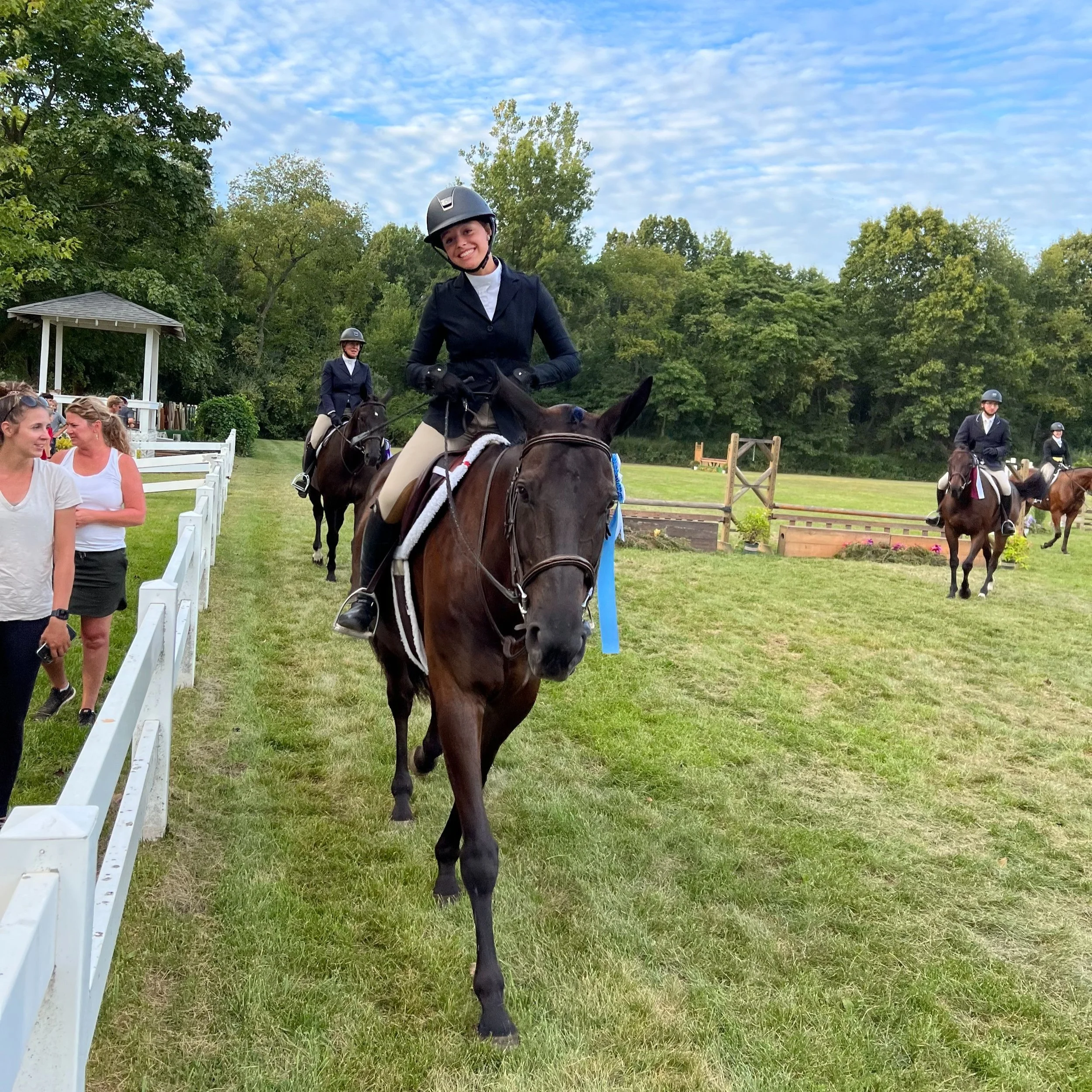 Young woman in riding attire and helmet smiling while riding a horse at an outdoor event with other riders and spectators around, greenery, and blue sky with clouds.