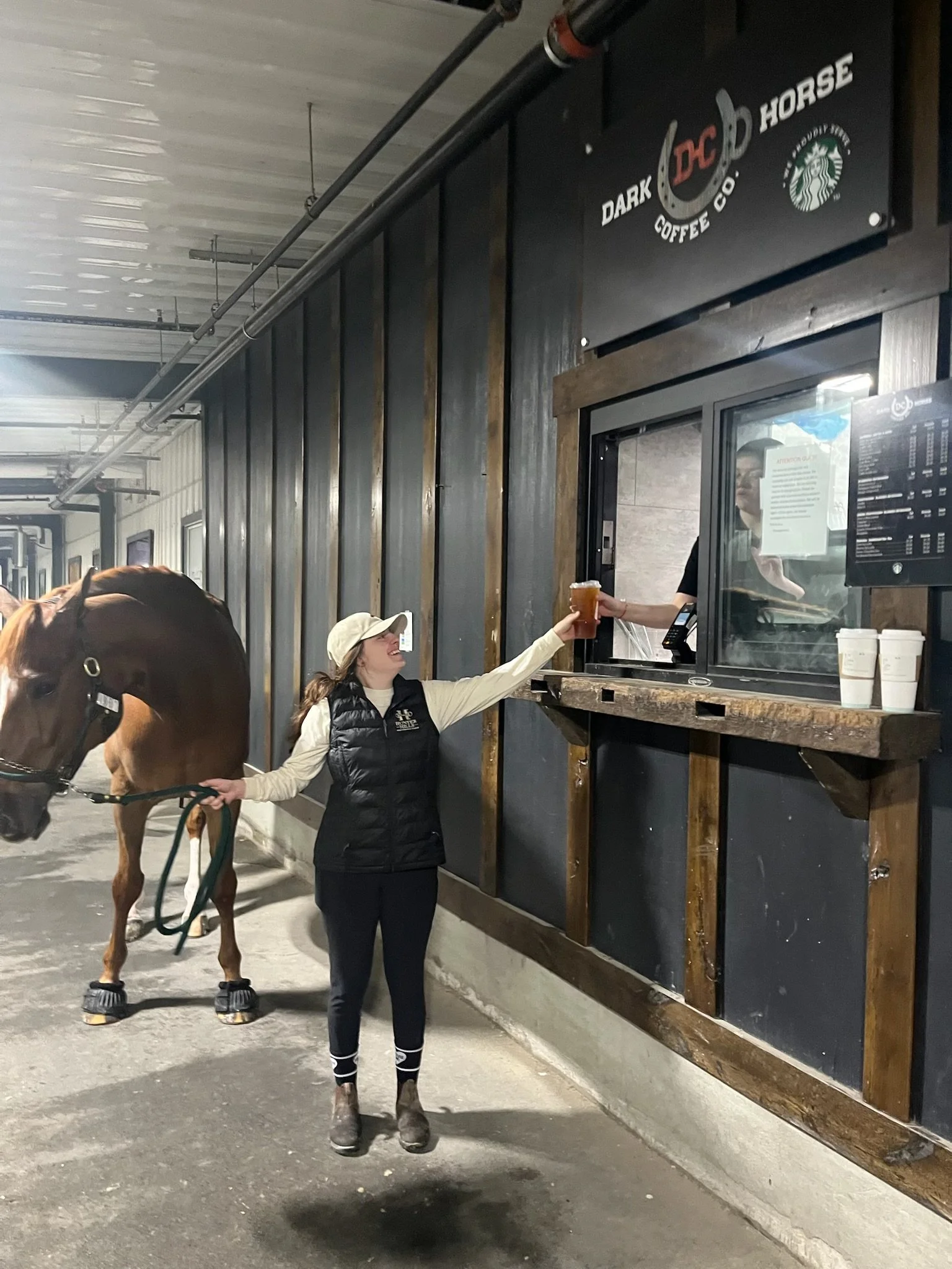 A female worker handing a cup of beer through a service window to a customer at a Red Horse Beer stand in a rustic, indoor setting with dark wooden accents and a sign reading "Dark Horse Coffee Co." and Starbucks logo.