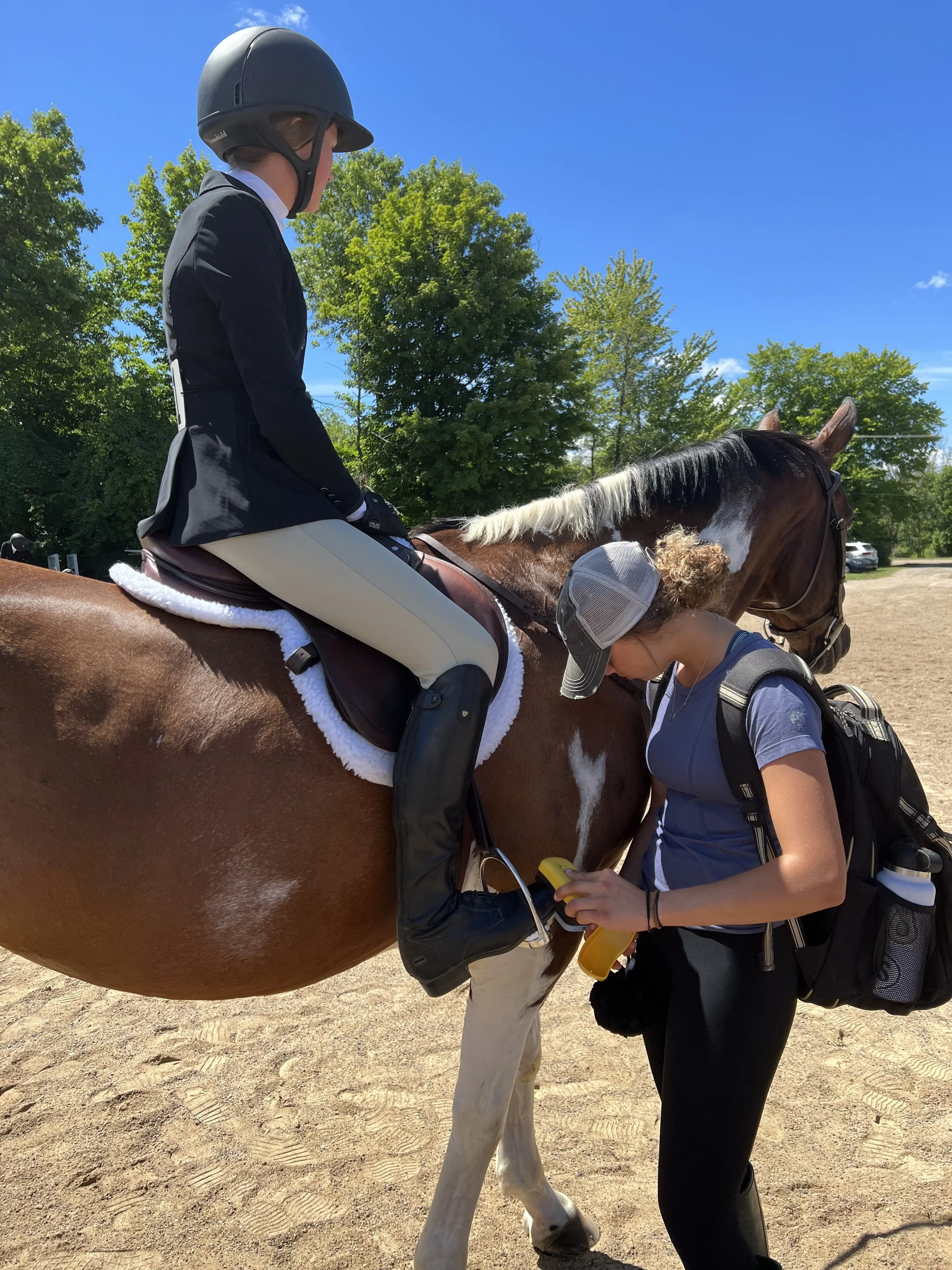 A woman riding a brown and white horse outdoors on a sunny day, with a person in a gray cap standing in front of the horse and holding a yellow device.