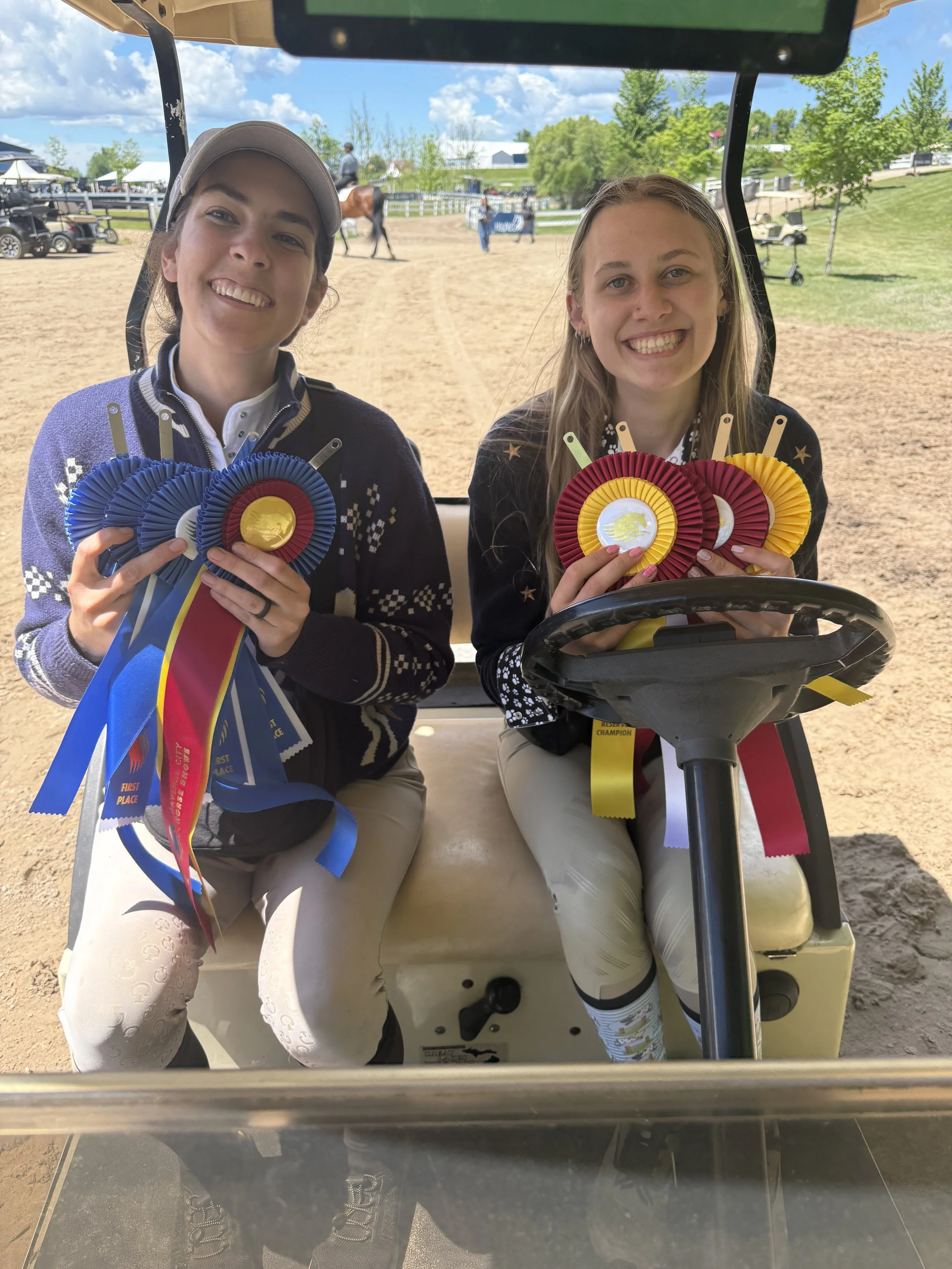 Two smiling women sitting in a golf cart, holding colorful award ribbons, at an outdoor golf course on a sunny day.