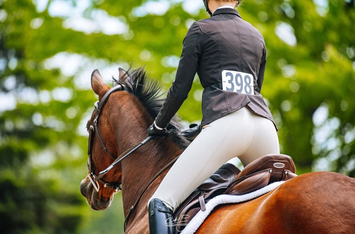 A rider on a brown horse during an equestrian event in a green outdoor setting. The rider is wearing a black jacket with a competitor number 398, beige riding pants, and black riding boots.