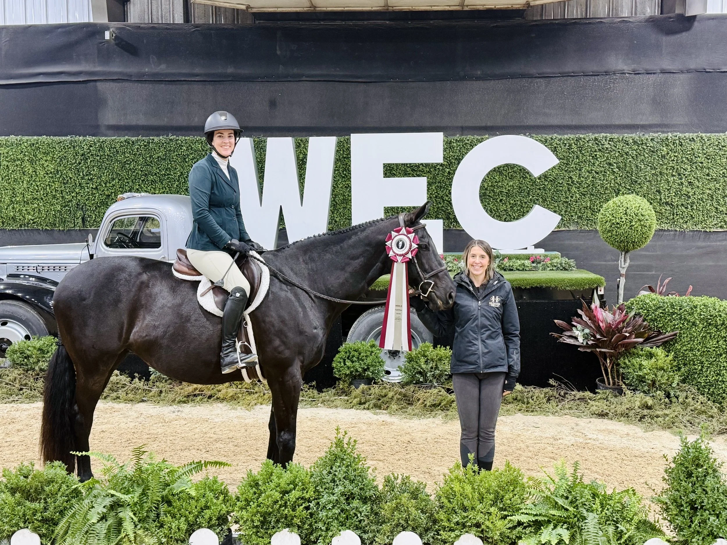 A woman riding a horse that is decorated with a ribbon, standing in front of a large sign that reads 'WEC' with a hedge background. Another woman stands beside the horse, smiling, in an outdoor setting with greenery and ornamental plants.
