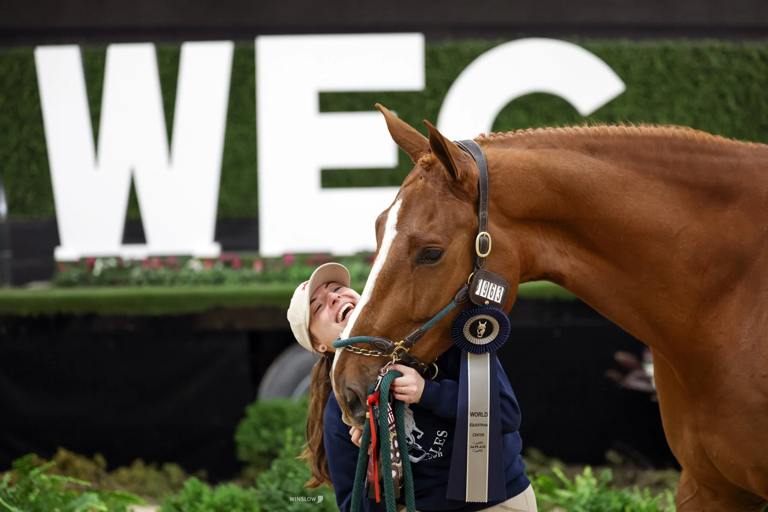 A young girl smiling as she hugs a brown horse with a white blaze on its face at a horse show. The girl is wearing a white cap and a dark jacket, and the horse has a black halter. In the background, there is a large white sign with 