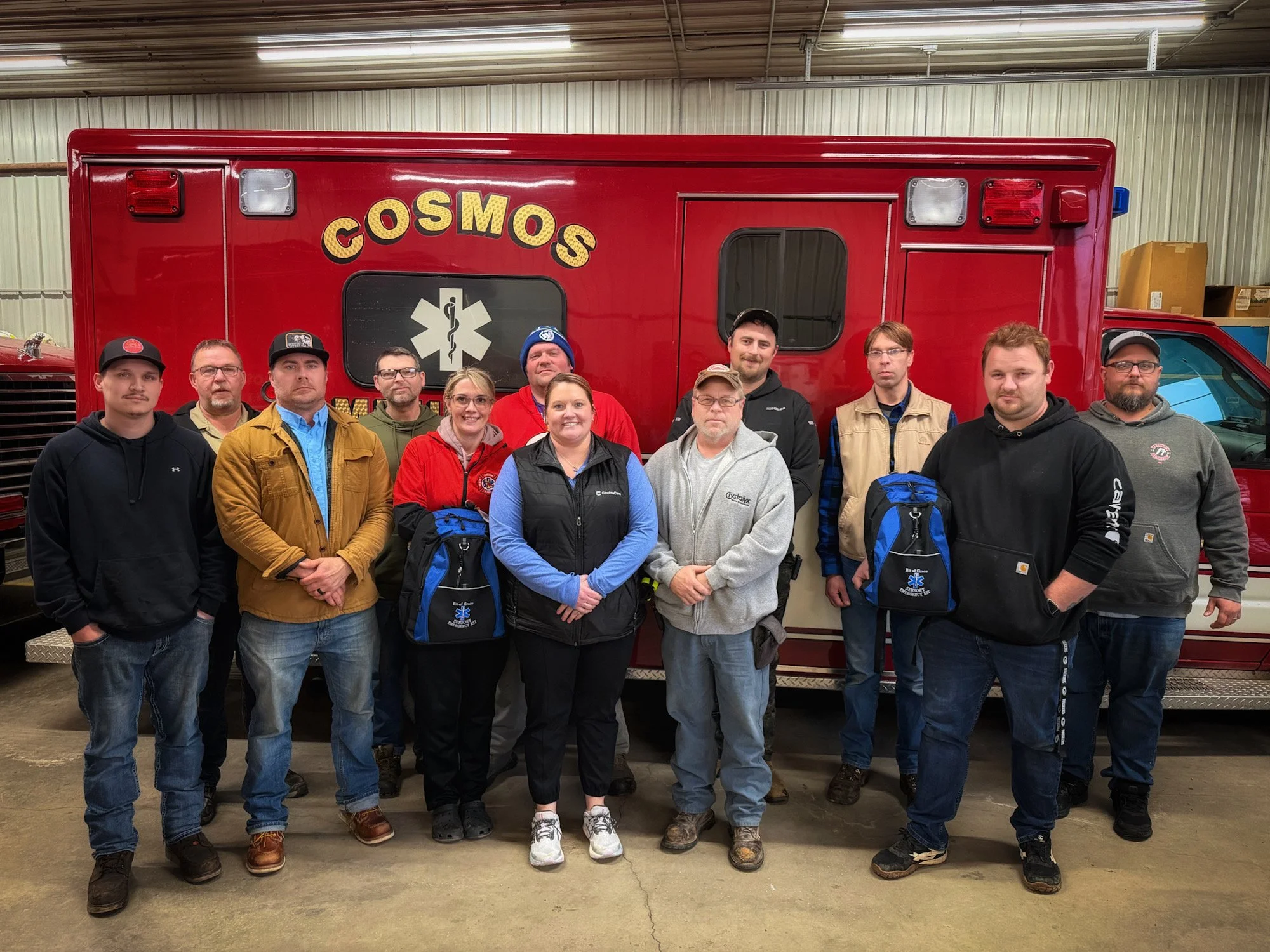 Group of eleven people standing in front of a red emergency vehicle with the word 'COSMOS' on it, inside a warehouse or garage.