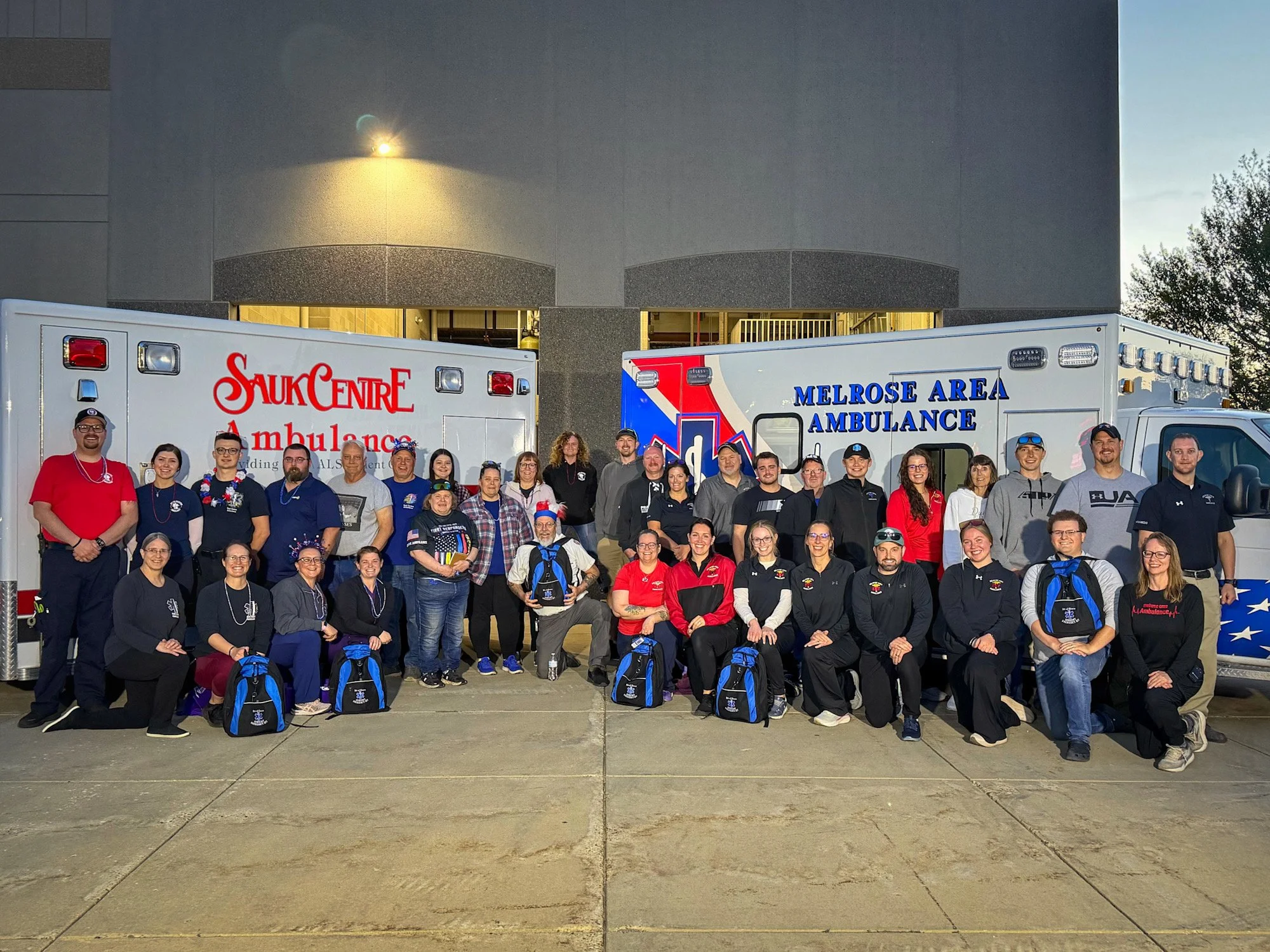Group of people standing in front of ambulances with medical staff and emergency responders, posing for a photo during daytime.