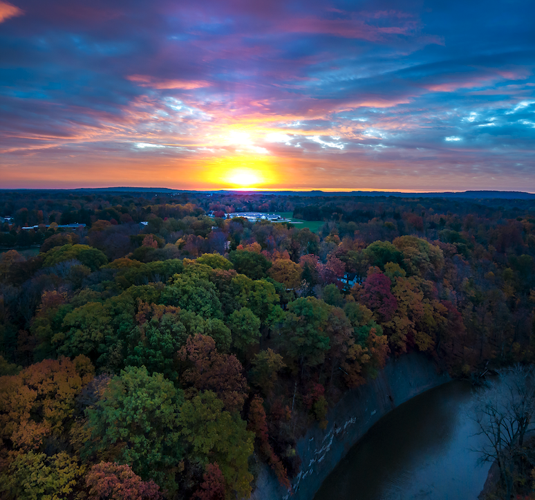 A vibrant sunset over a landscape with a dense forest showing fall foliage, a river curving along the edge of the trees, and the sky filled with colorful clouds.