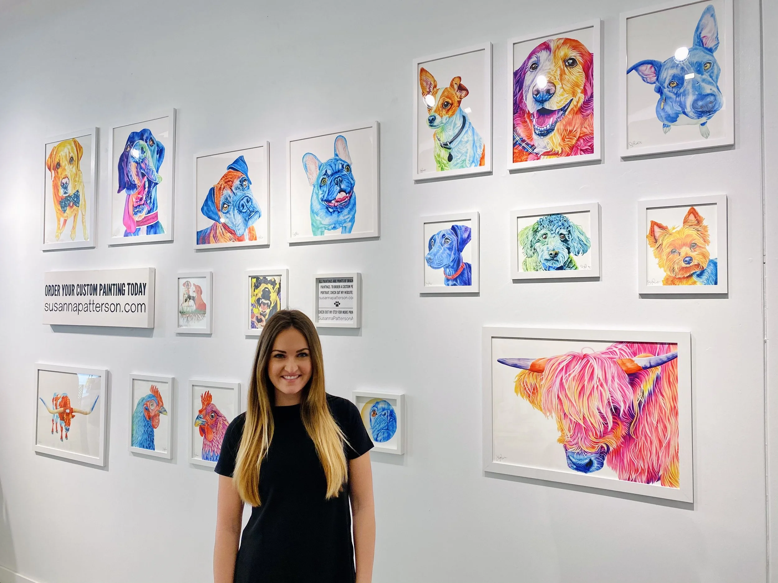 Artist Susanna Patterson smiling before a gallery wall of vibrant, rainbow-hued animal portrait prints in white frames.