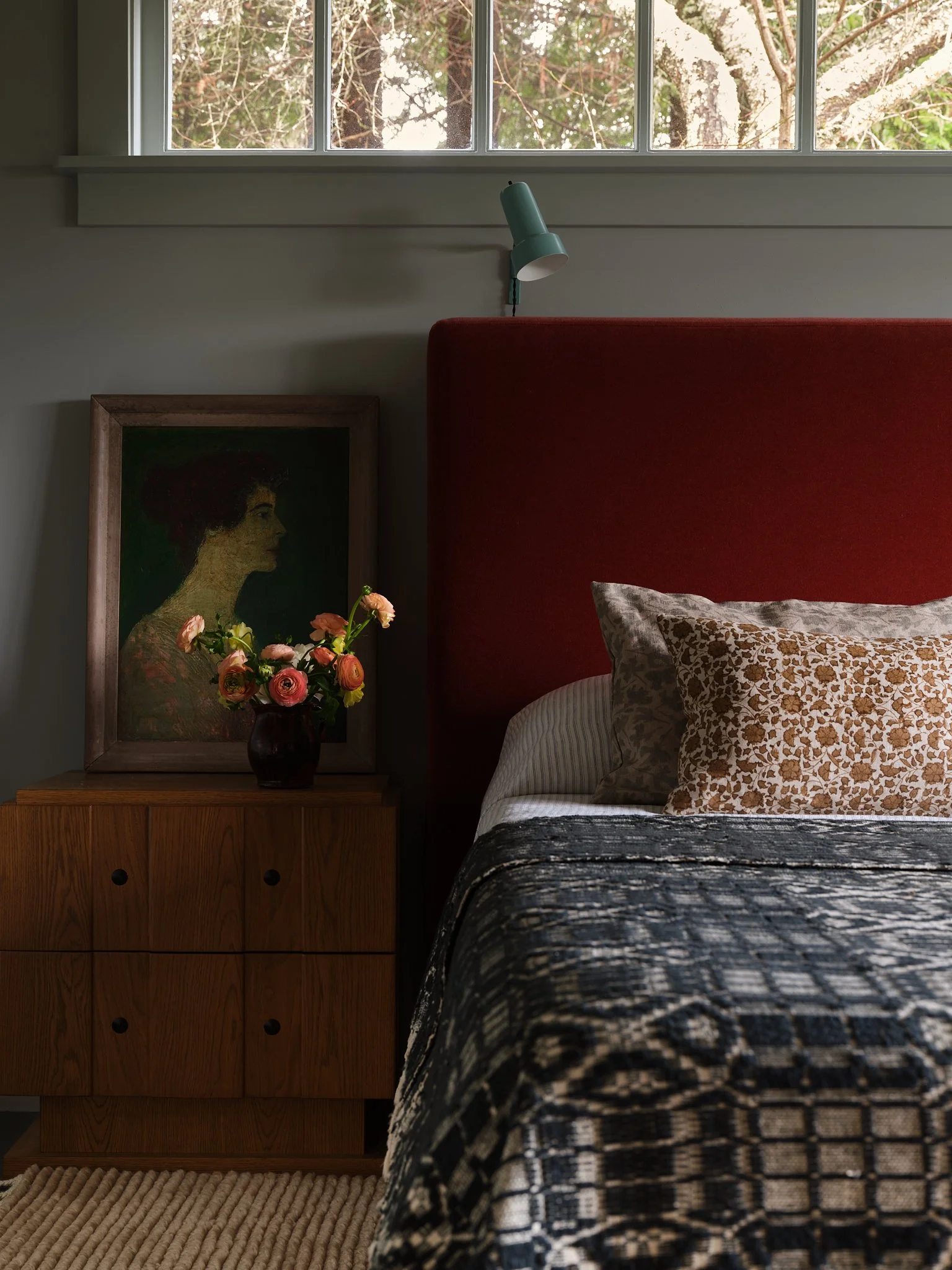 A bedroom with a red upholstered headboard, a wooden nightstand holding a framed portrait and a vase of pink flowers, and a window showing trees outside.