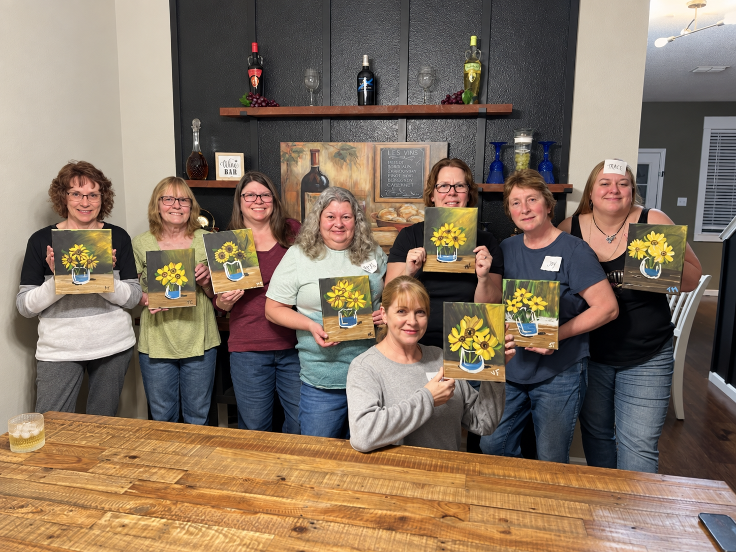 A group of nine women standing indoors, each holding a painting of yellow flowers in a blue vase. They are smiling and posing for the photo, with a black wall featuring shelves, decorations, and a framed painting of wine bottles in the background.