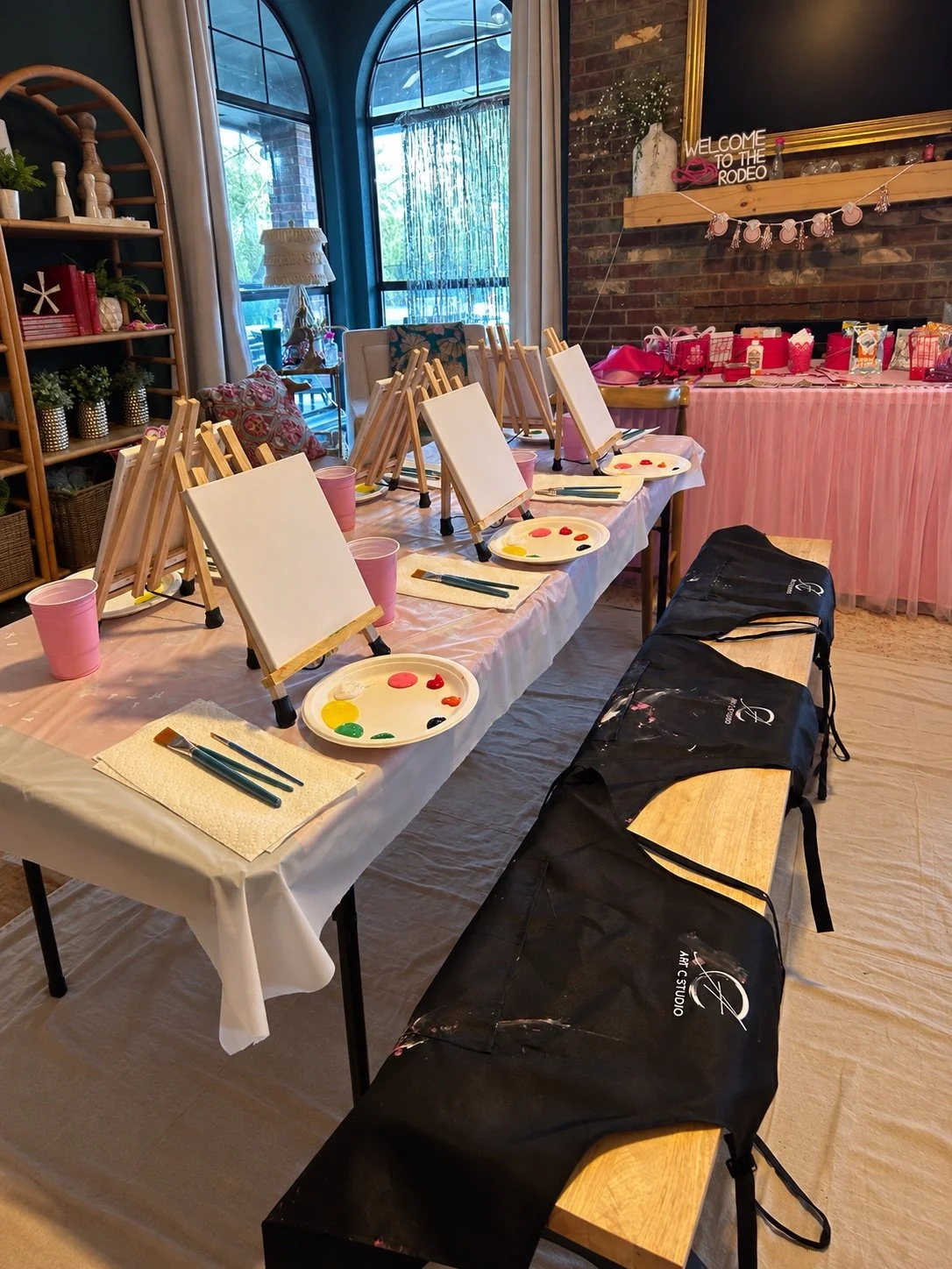 Indoor room set up for a painting activity with small canvases on easels, paint palettes, brushes, pink cups, and black aprons on a wooden bench. There are decorations including a 'Welcome to the Rodeo' sign, pink garland, and various gift bags on a pink tablecloth at a table with a pink and white theme.