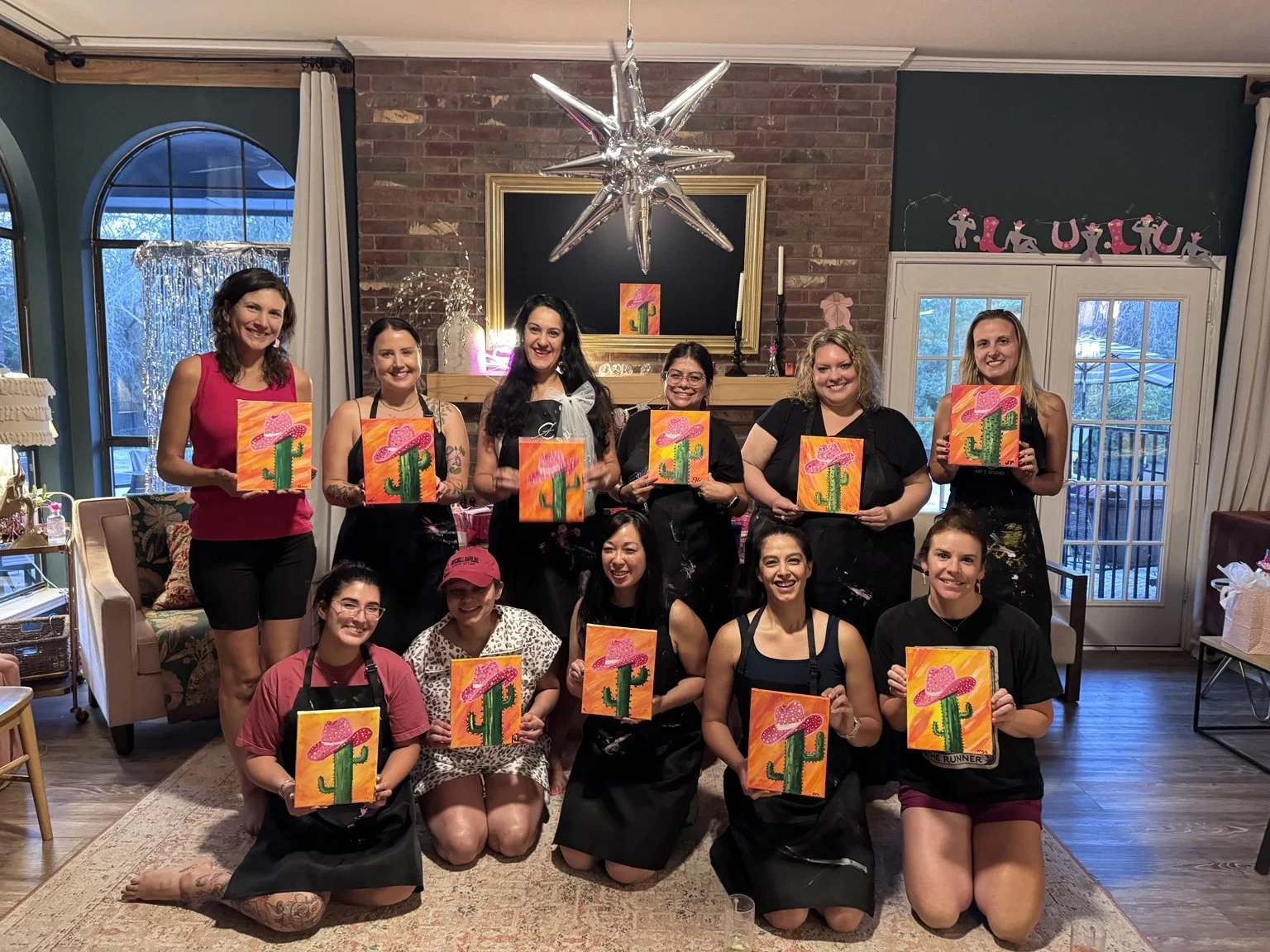 A group of ten women in a living room holding up paintings of a cactus with a pink sombrero hat, with some sitting and others standing, celebrating a crafting event during daylight hours.