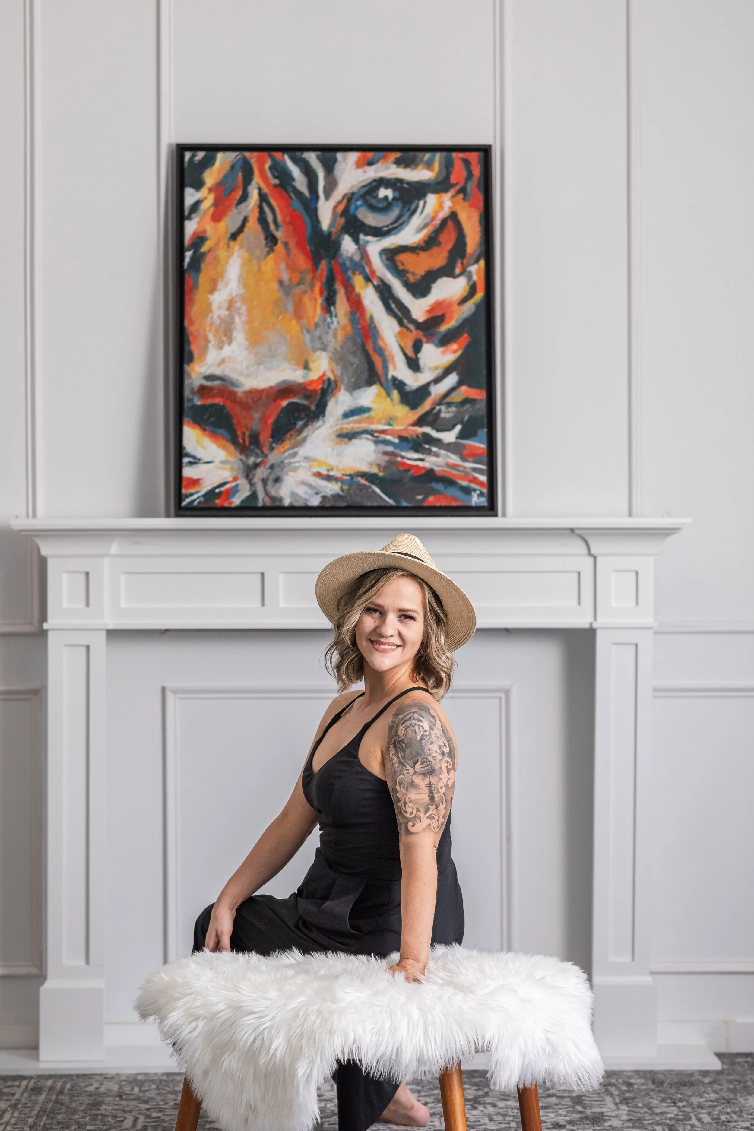 A woman with blonde hair, wearing a tan hat and a black dress, smiling and sitting on a white furry stool in a room with a white fireplace and a colorful tiger artwork on the wall behind her.