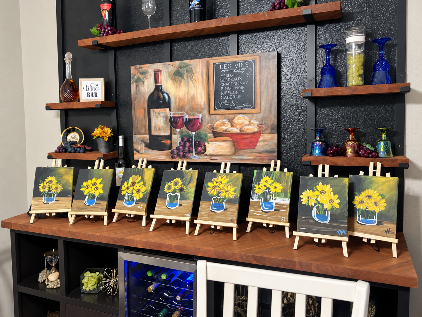 A display of eight small canvas paintings of yellow sunflowers in blue vases on easels arranged on a wooden table. The background features a large still life painting of a wine bottle, two glasses of red wine, grapes, bread, and a chalkboard menu listing various wines. Shelves above hold decorative bottles, grapes, a glass, a clock, and small art pieces.