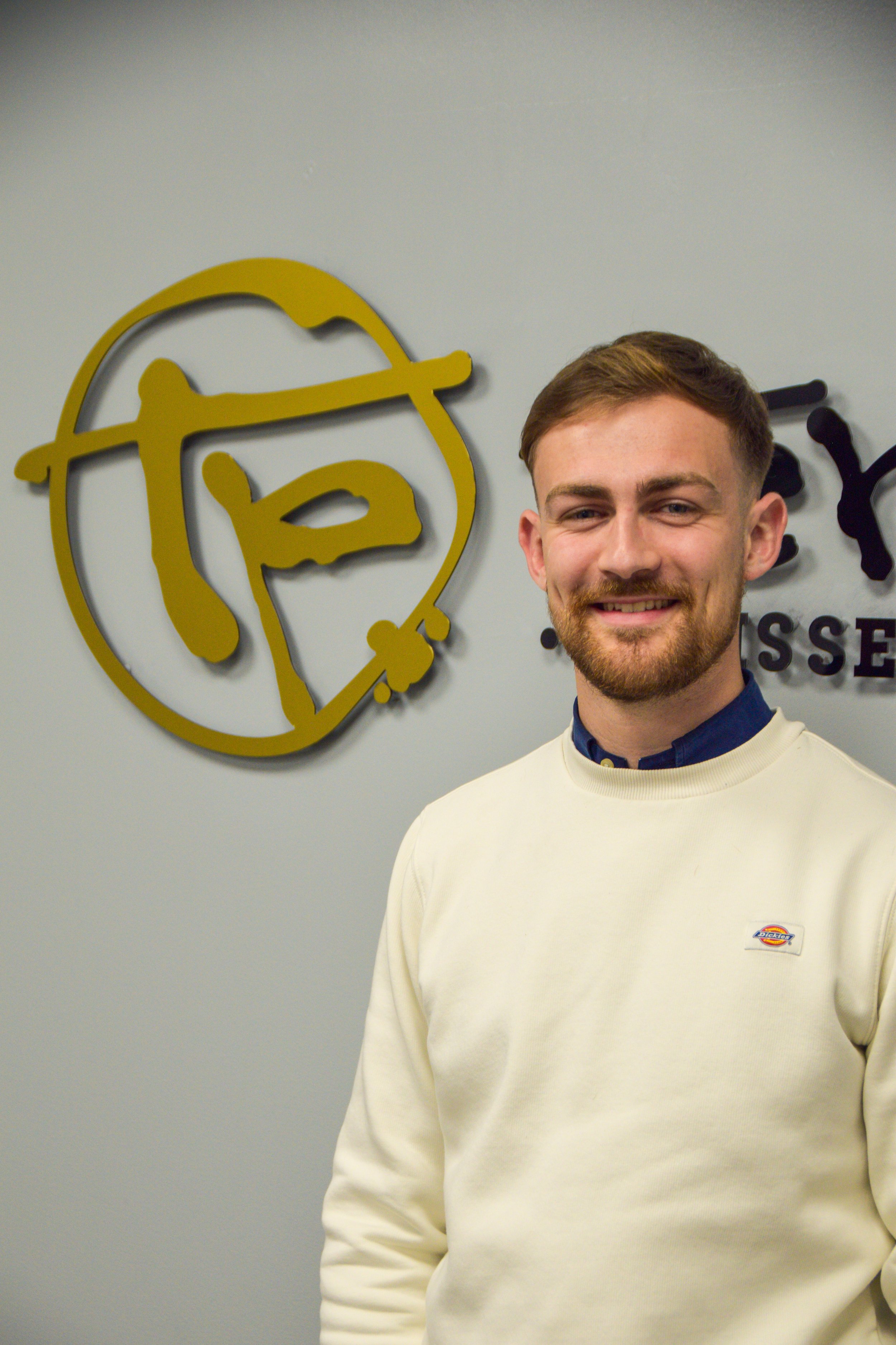 Young man in dress shirt holding papers, standing in front of a presentation slide, in a classroom or conference room setting.