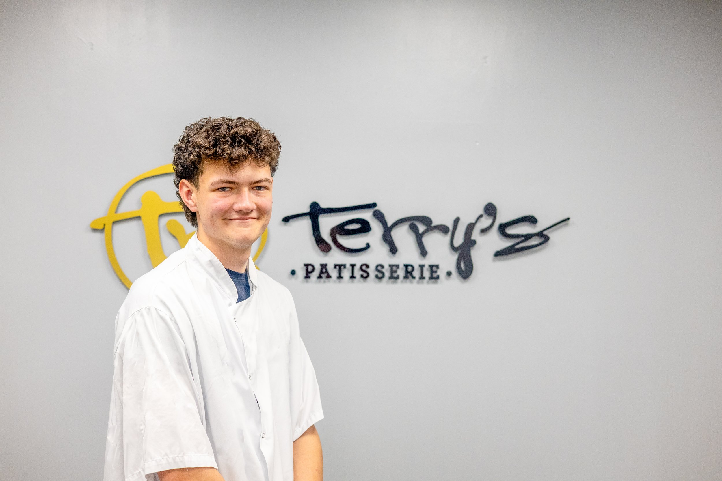 Young man in chef uniform with black apron standing in front of a wall with 'Terry's Patisserie' sign.