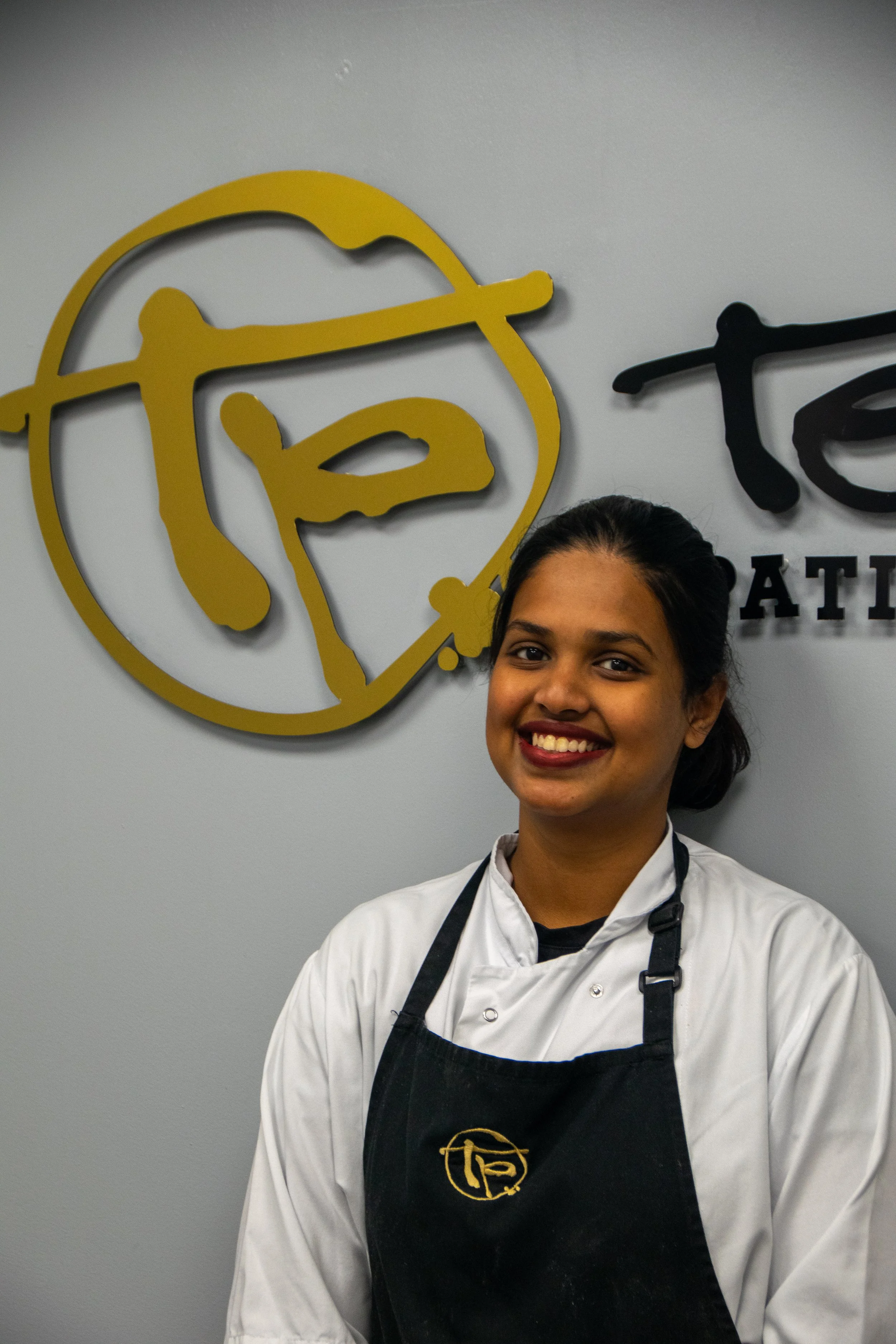 A woman in a chef's coat standing in front of a gray wall with a sign that reads 'Terry's Patisserie'. She has her hands on her hips and is smiling.