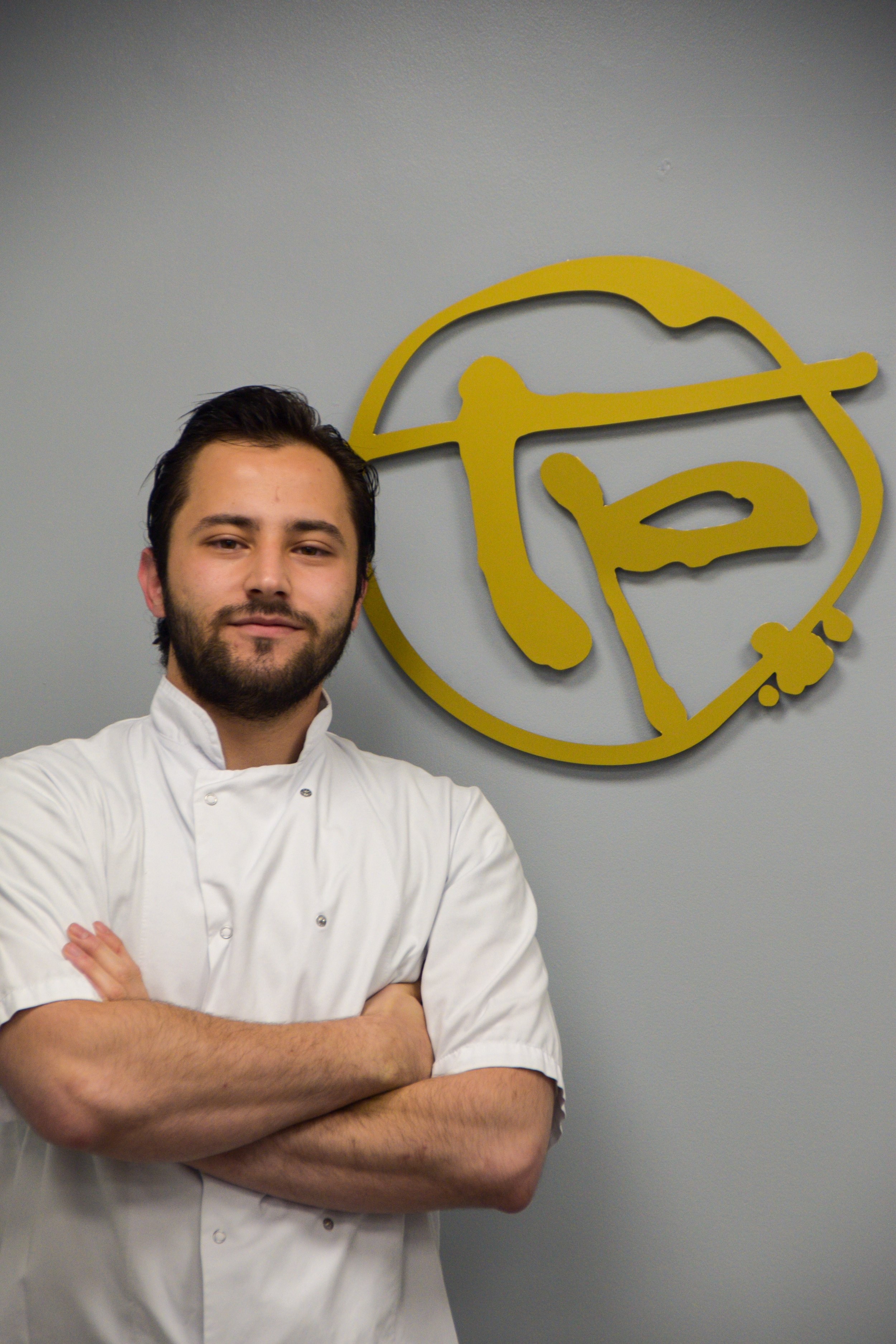 A man wearing glasses and a white chef's coat with a black apron stands with his arms crossed in front of a black wall with partially visible white and yellow text.