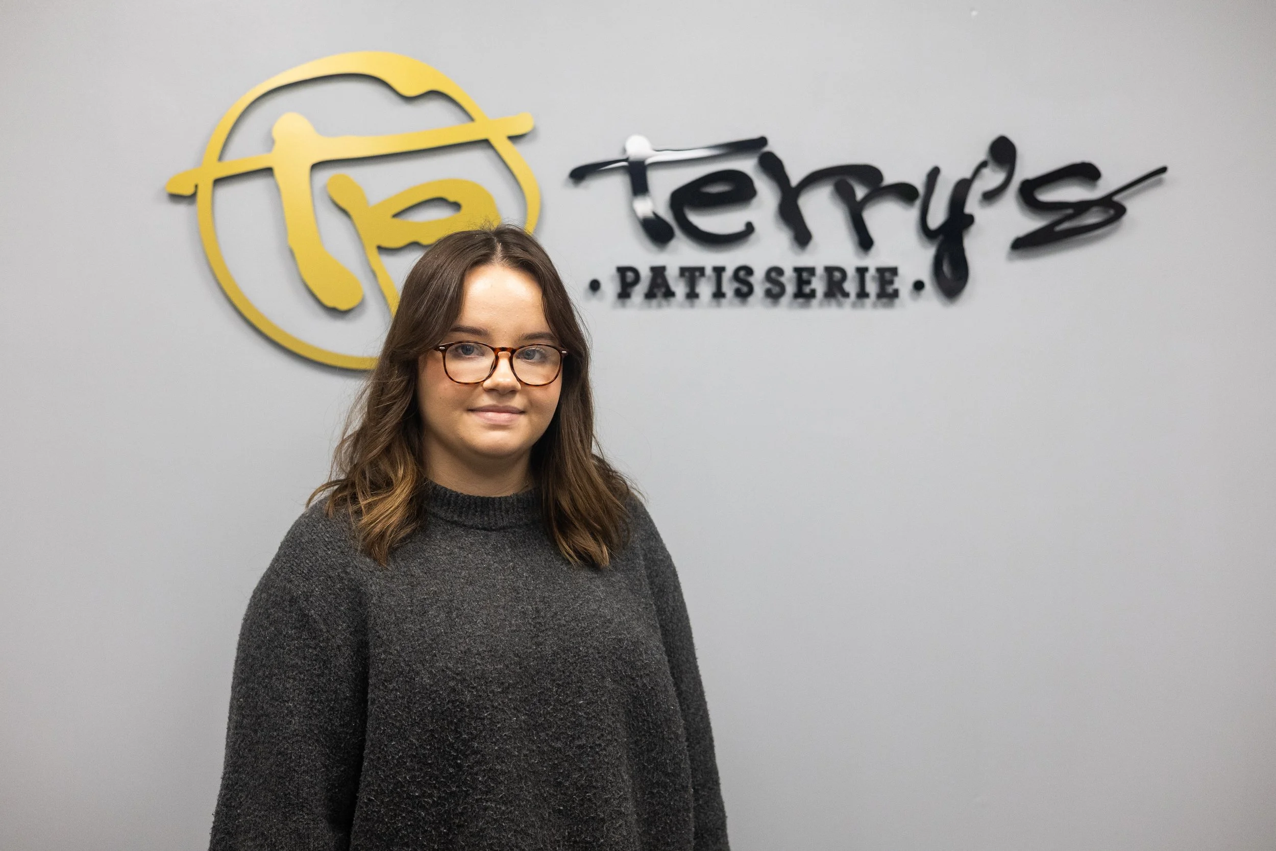 A young woman with glasses and long brown hair standing in front of a wall with the logo of Tery's Patisserie. She is wearing a black and white striped sweater and blue jeans, smiling at the camera.